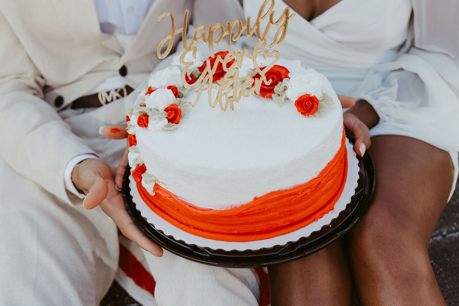 Couple holding a white and orange wedding cake topped with a Happily Ever After gold topper.