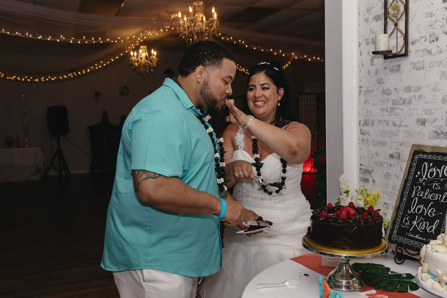 Bride playfully feeds groom wedding cake during fun reception celebration with string lights.