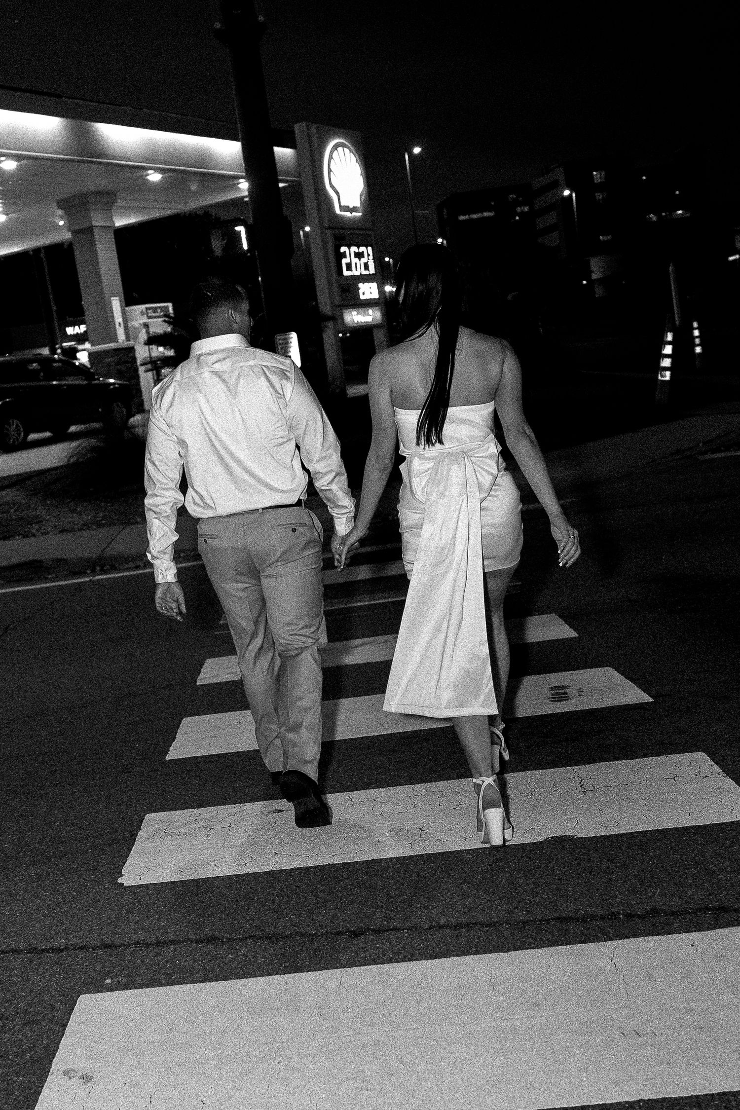 Black and white photo of couple holding hands crossing street at night near Shell gas station, woman in white dress.