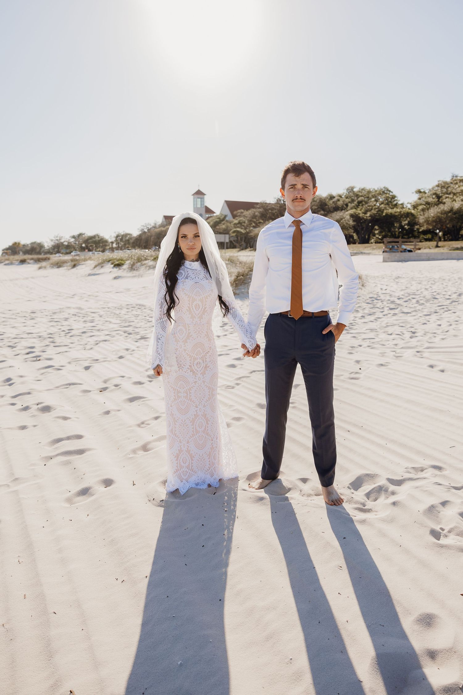 Bride in lace gown and veiled groom hold hands on bright sandy beach at sunset.
