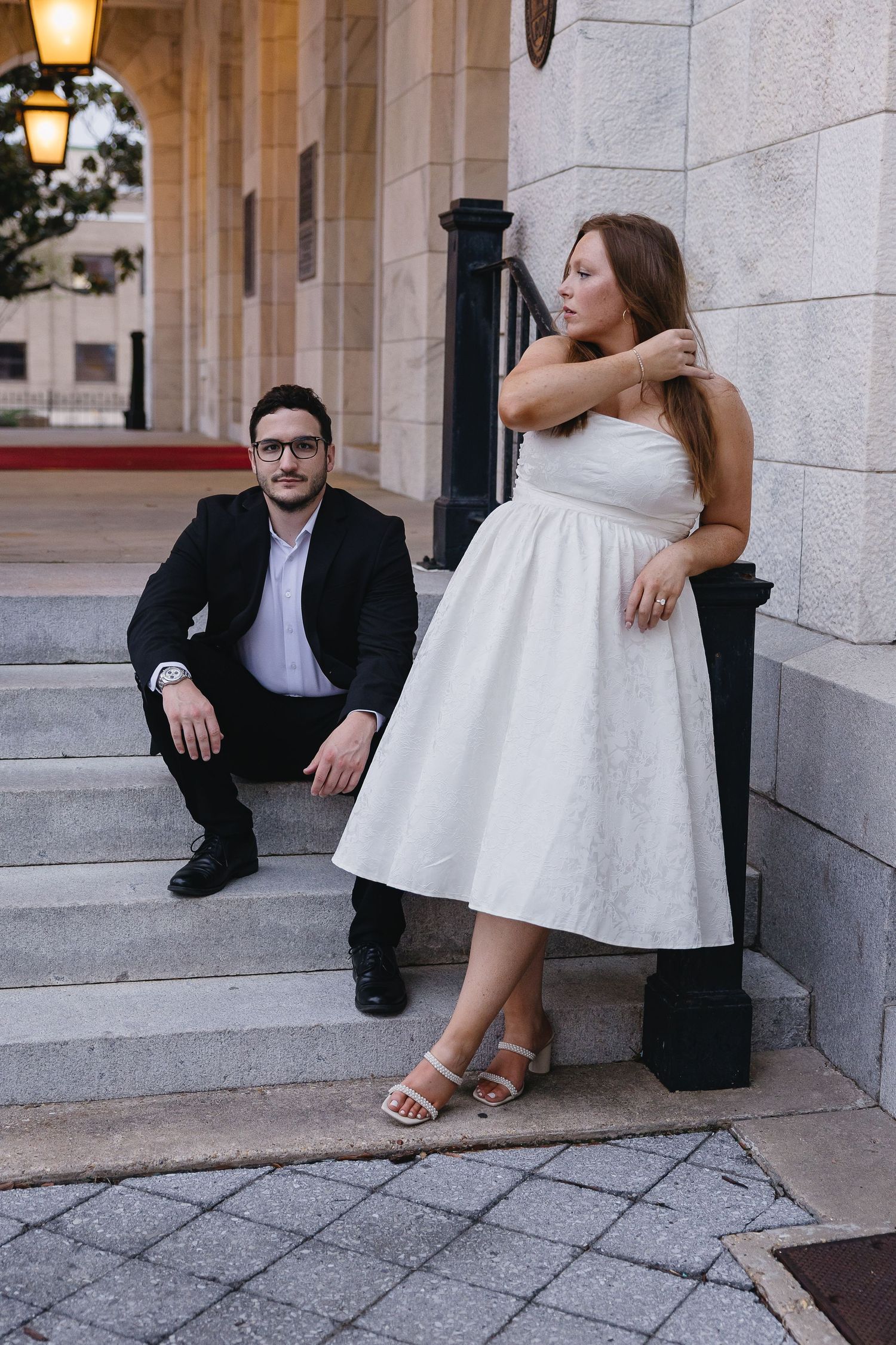 Bride in white dress leaning on railing while groom sits on steps gazing up at her.