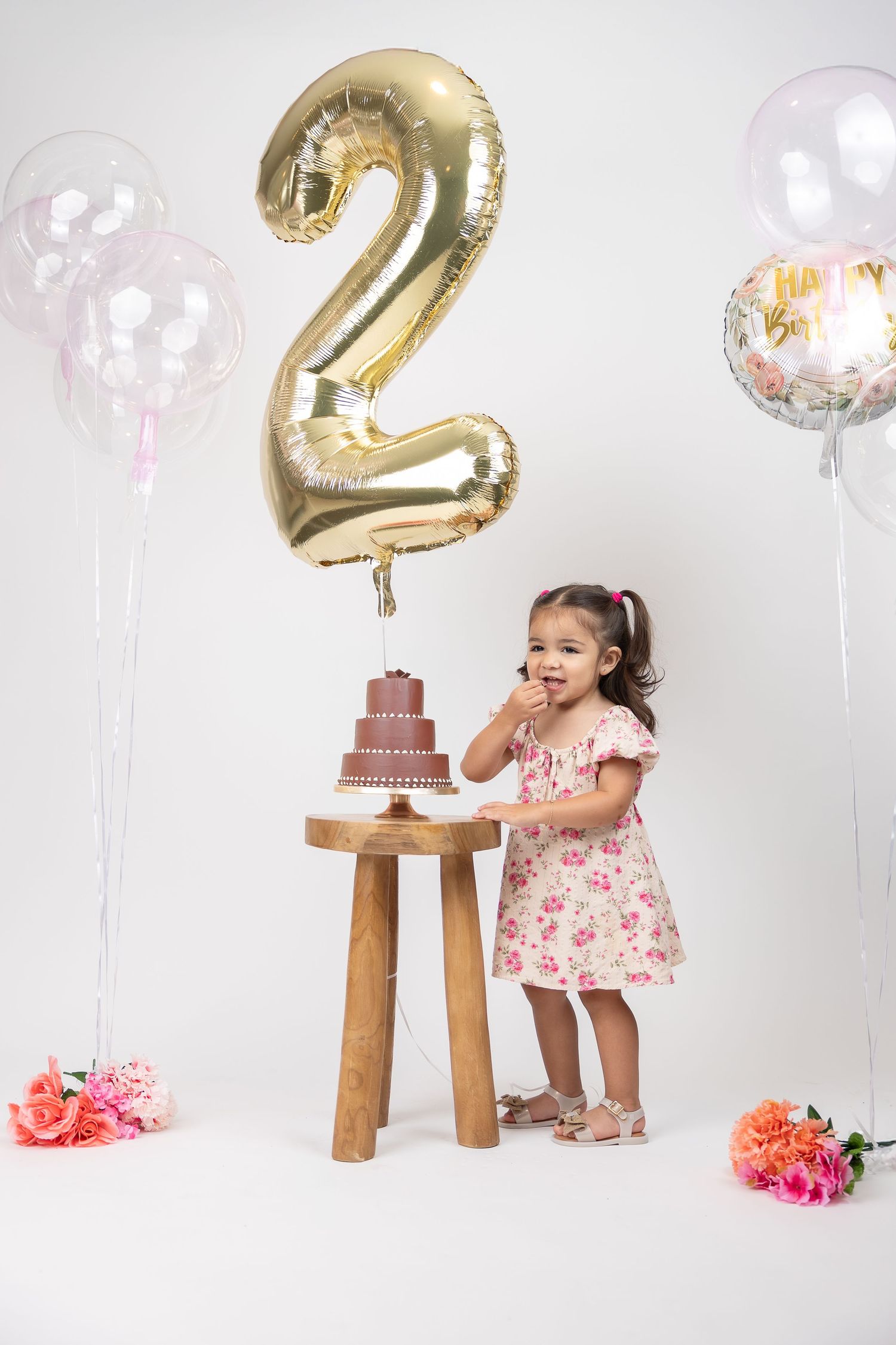 Toddler girl in floral dress celebrating 2nd birthday with cake and gold number balloon.