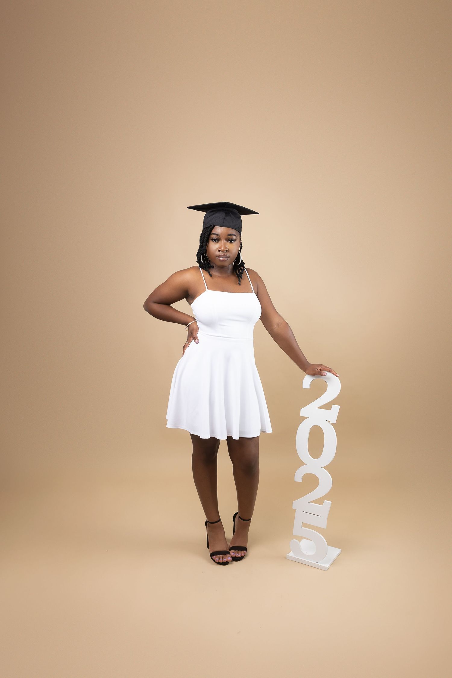 A young Black woman in a white dress and graduation cap poses with a 2025 sign on beige background.