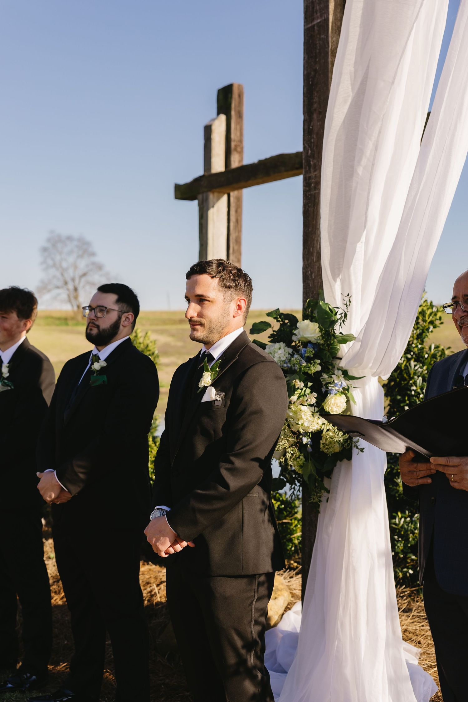 Groom in black suit standing at wedding altar with wooden cross and white floral draping outdoors.