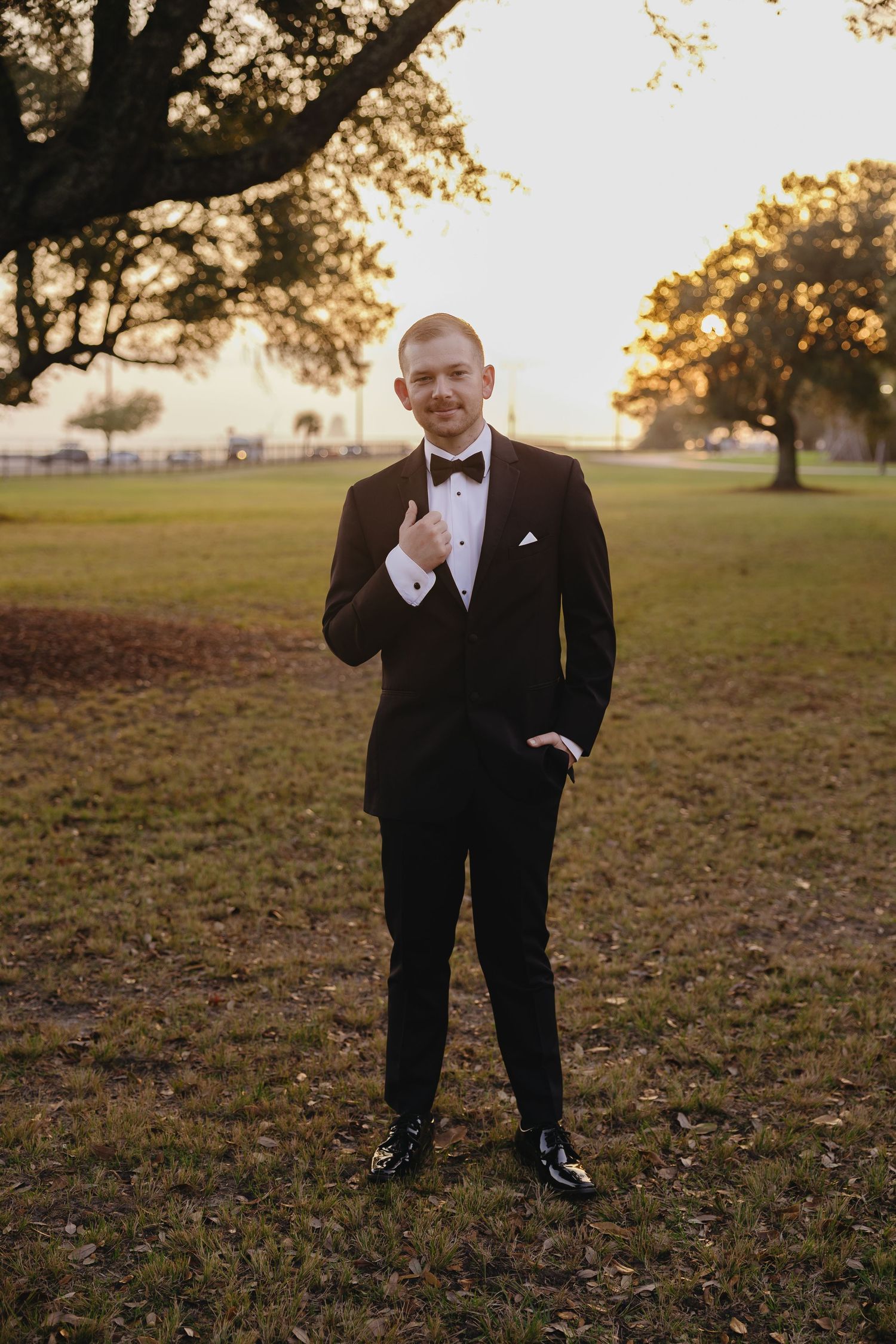 Dapper groom in black tuxedo and bow tie poses confidently in a sunlit park at golden hour.