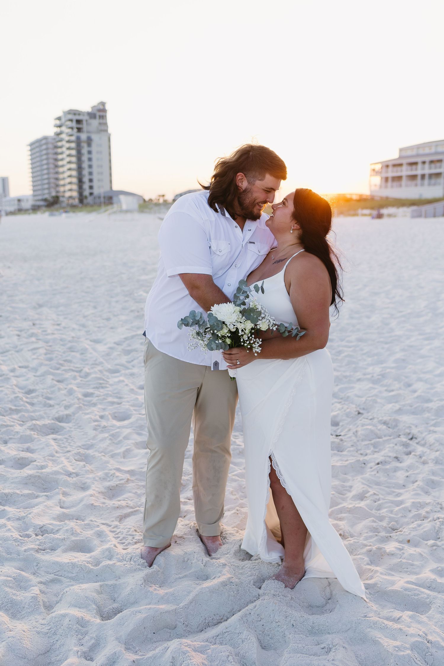 A couple shares a romantic kiss on a sandy beach at sunset, bride holding a white floral bouquet.