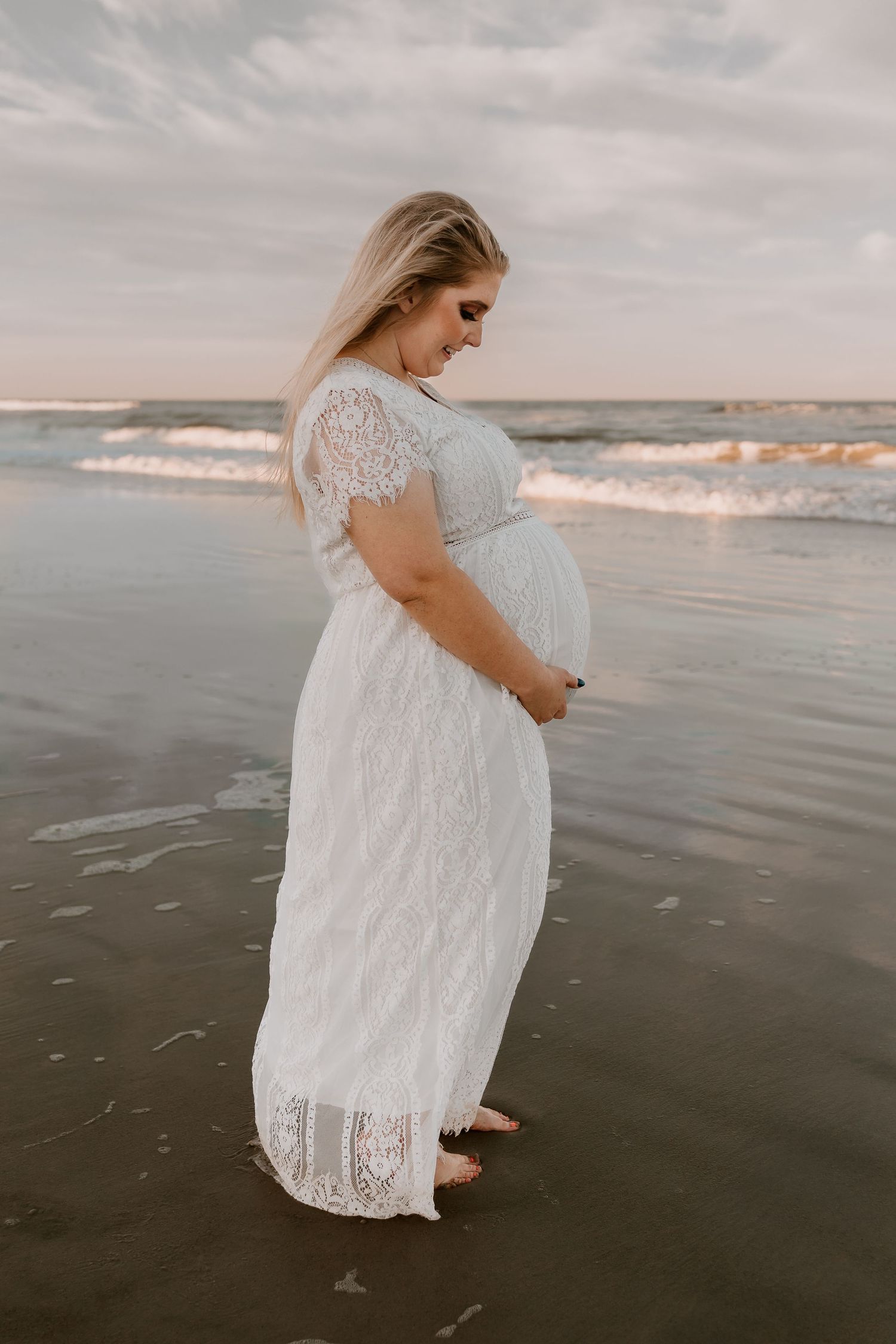 Pregnant woman in white lace dress standing at ocean's edge on beach, holding belly, waves behind her.