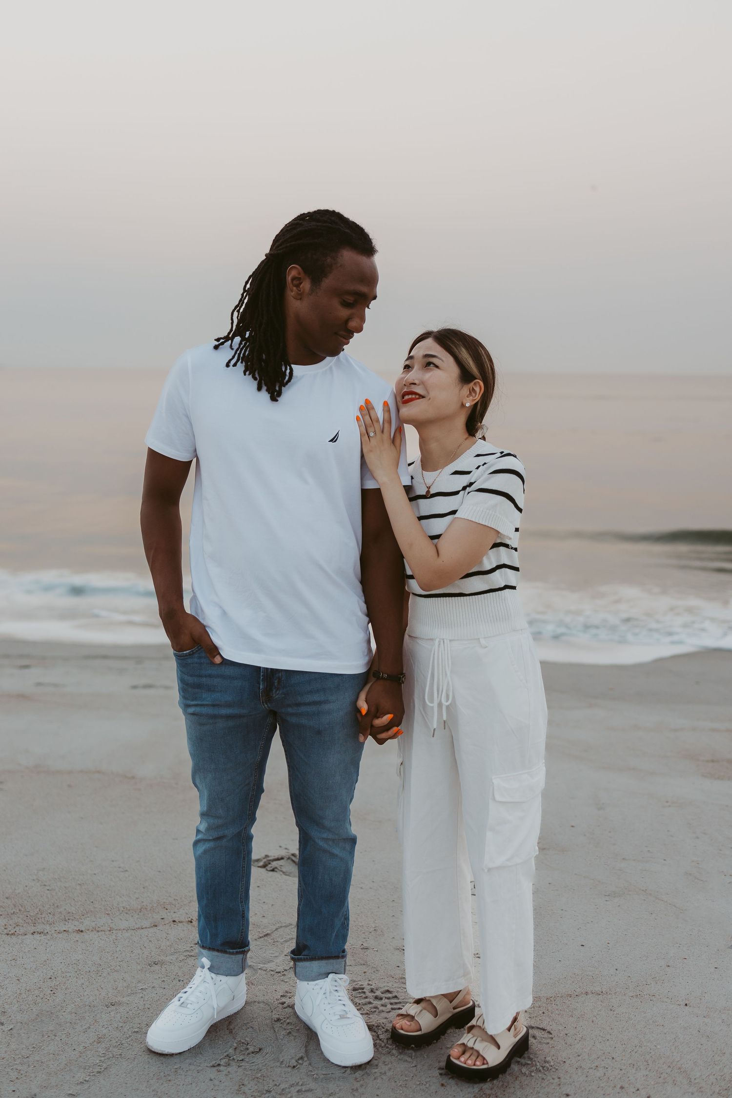 Interracial couple holding hands and gazing at each other on a beach at dusk.