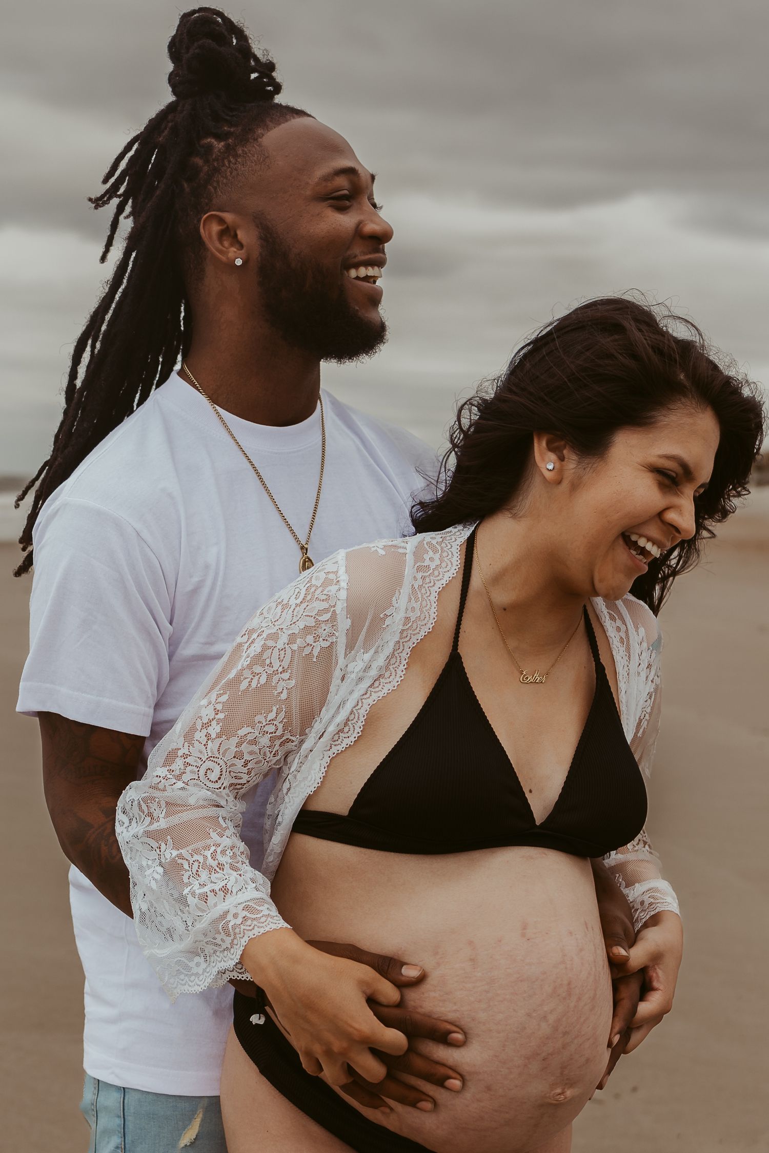Happy interracial couple laughing on beach, man holding pregnant womans baby bump outdoors.