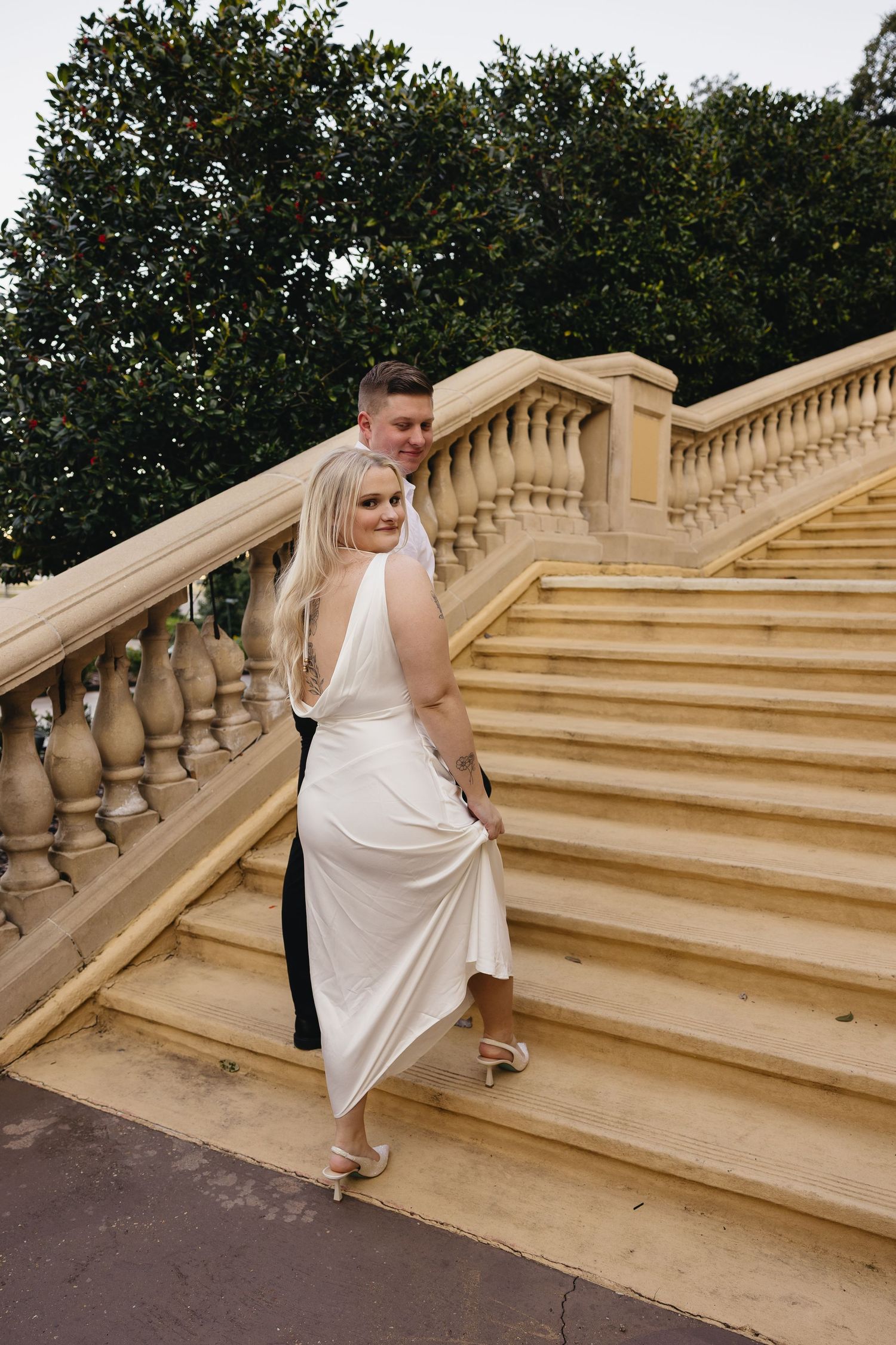 Couple in wedding attire sharing romantic moment on grand stone staircase with ornate balusters outdoors.