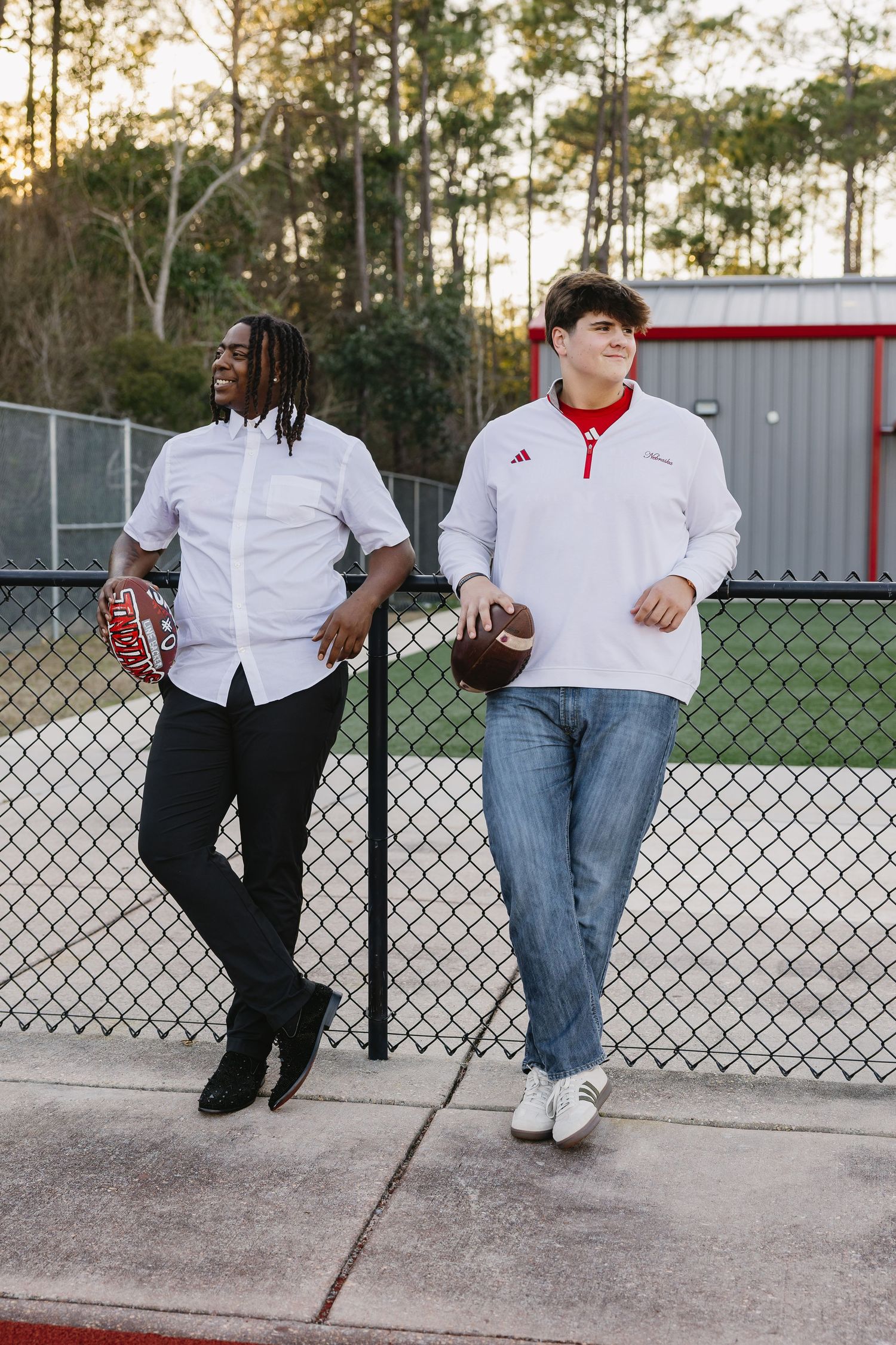 Two high school senior football players leaning against chain-link fence holding footballs at sunset.