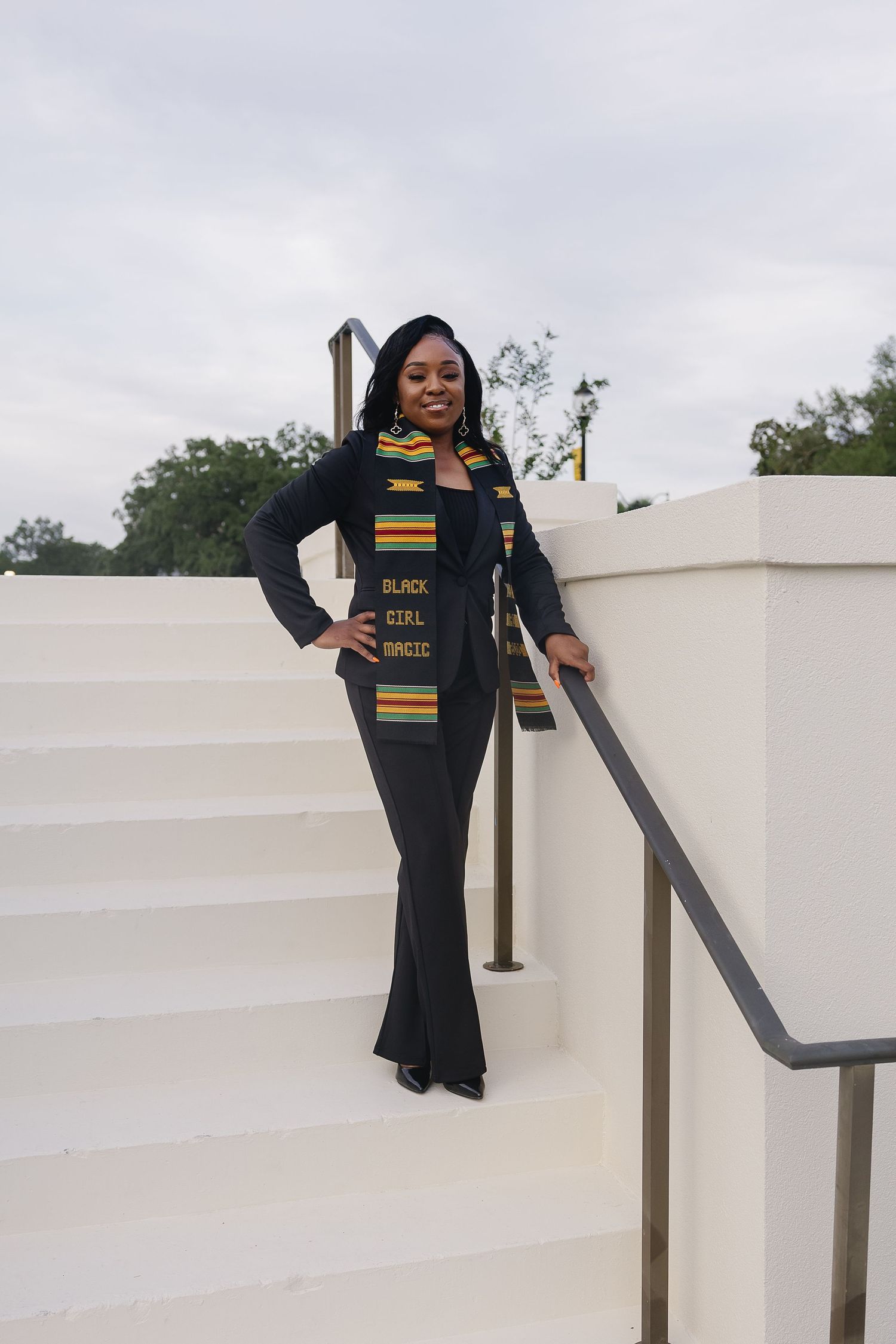 A confident graduate in black flare pants and Black Girl Magic kente stole poses on white steps.