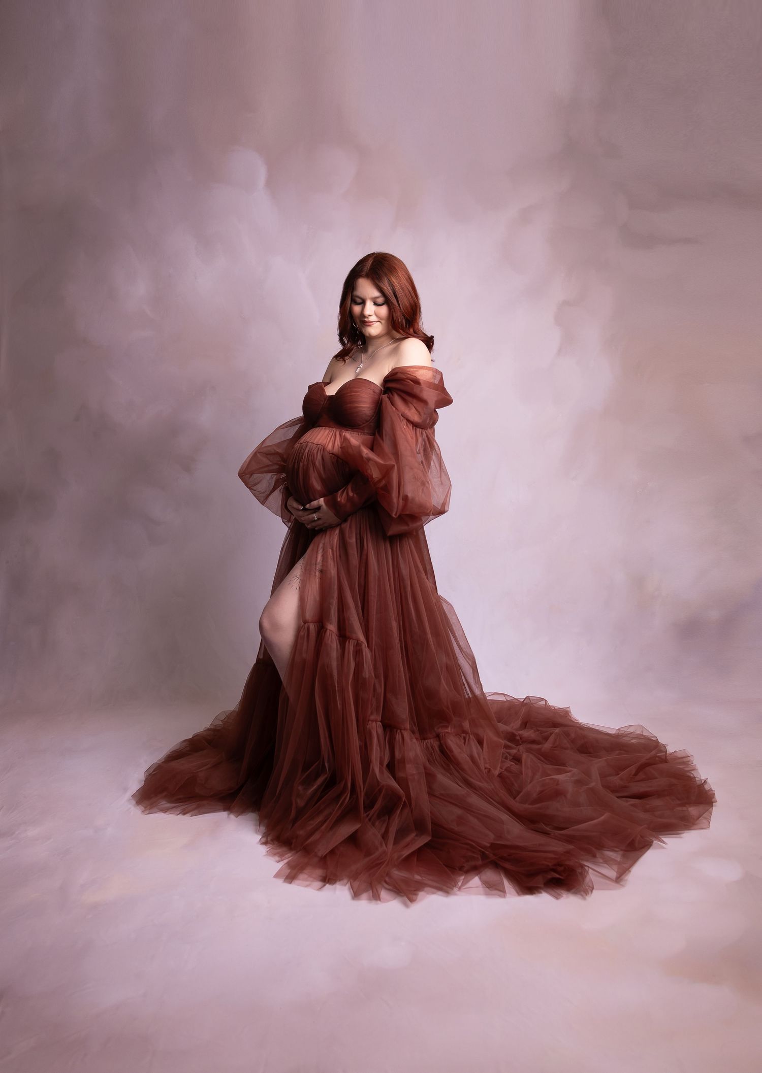 Pregnant redhead woman in off-shoulder rust tulle gown poses gracefully on pink backdrop.