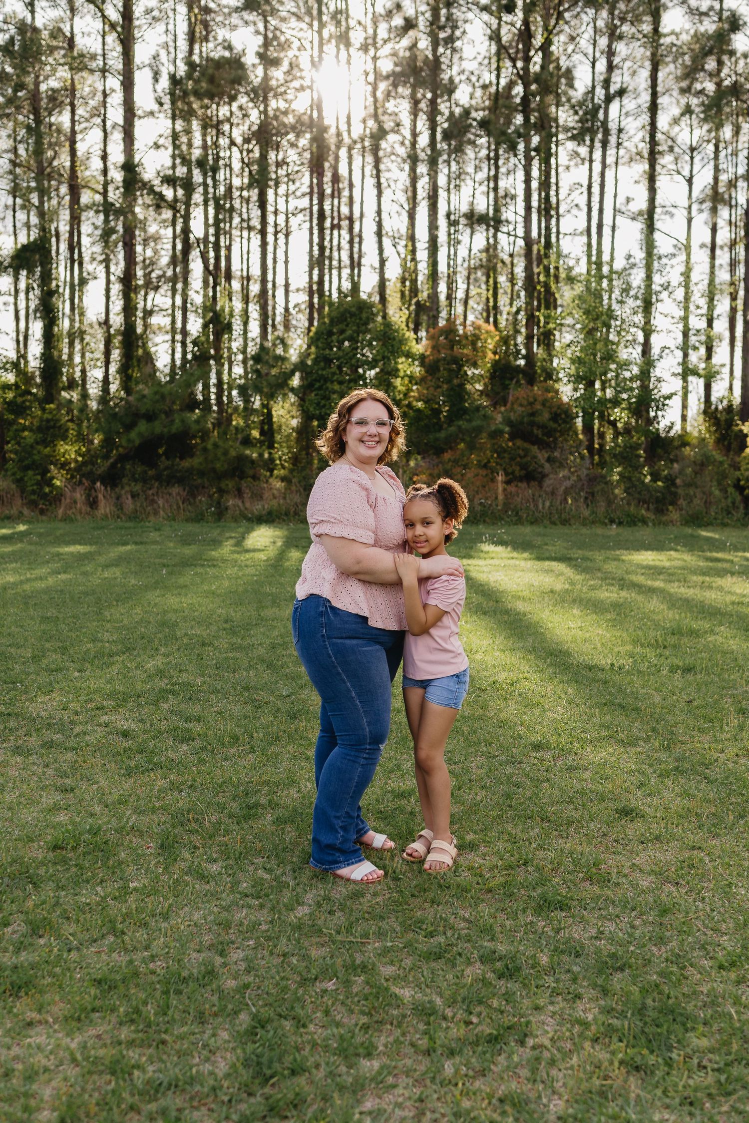 A mother and daughter in matching pink tops embrace in a sunny green field surrounded by tall pine trees.