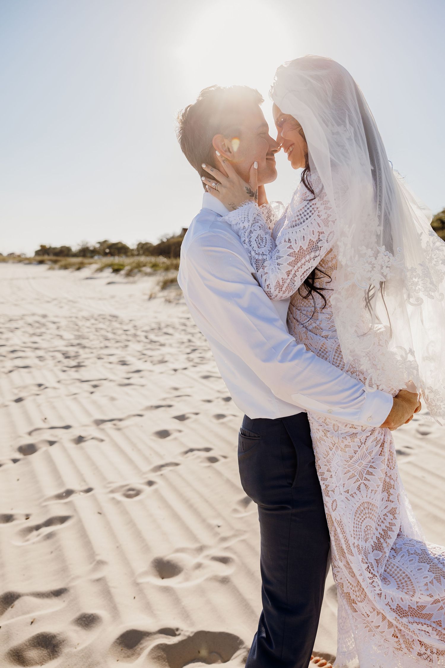 Bride and groom embracing on a sunlit sandy beach, bride wearing lace gown and veil.