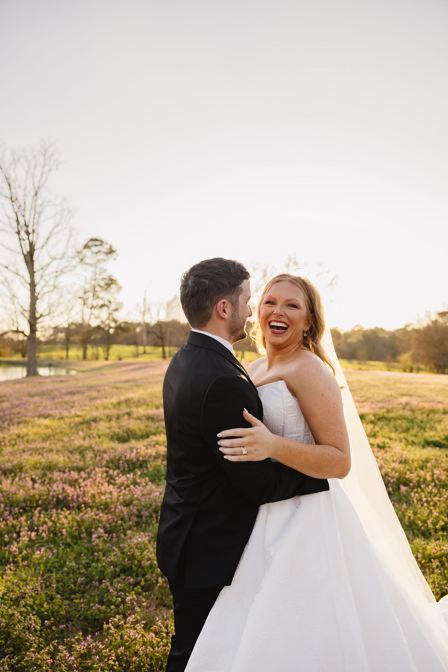 Laughing bride in white gown embracing groom in black suit in blooming meadow at golden hour.