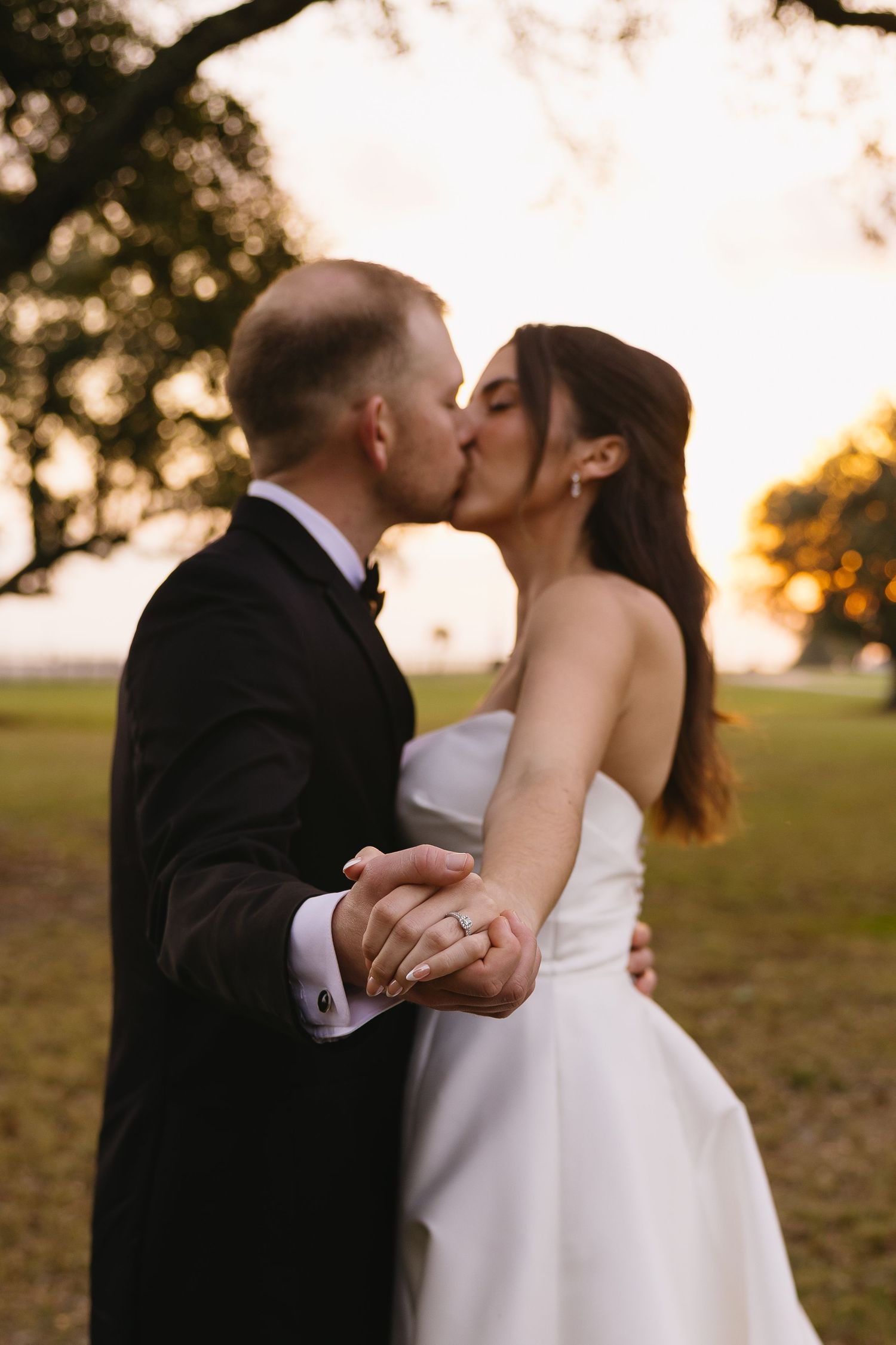Bride and groom share a romantic kiss at sunset while holding hands outdoors during wedding portraits.