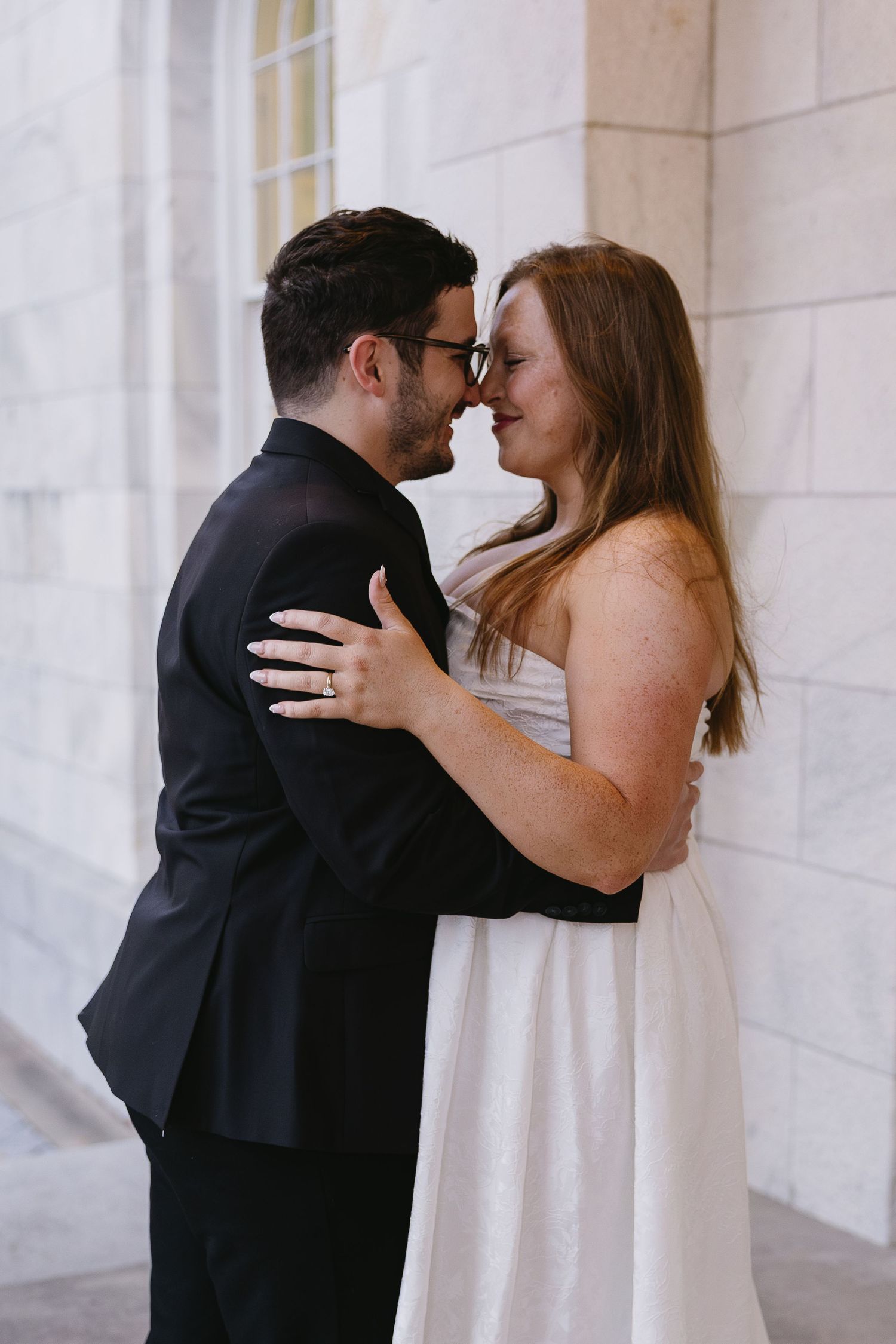 Couple embracing and touching noses in front of stone columns, bride in white strapless dress and groom in black suit.