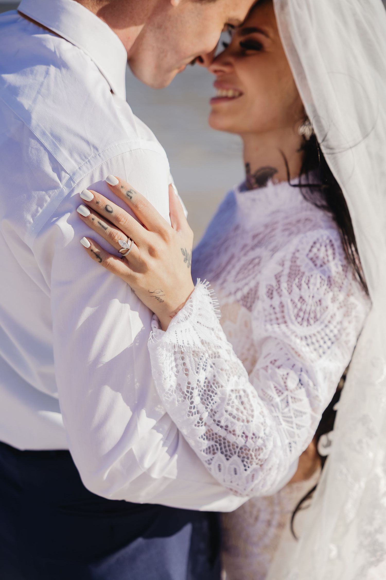 Bride in lace dress with tattooed hands embraces groom closely during beach wedding.