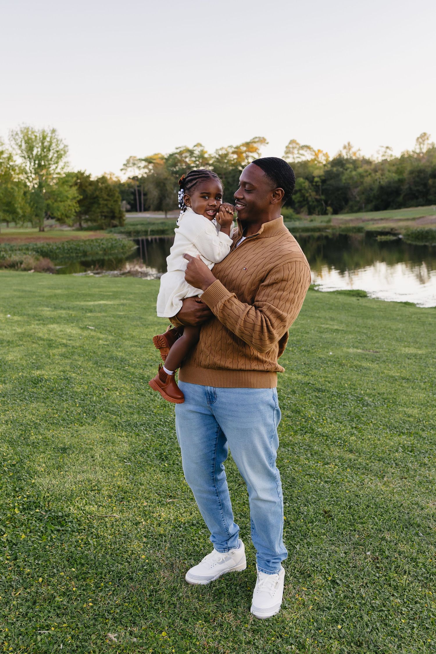 Happy Black father holding and gazing at young daughter in white dress outdoors near a scenic pond at dusk.