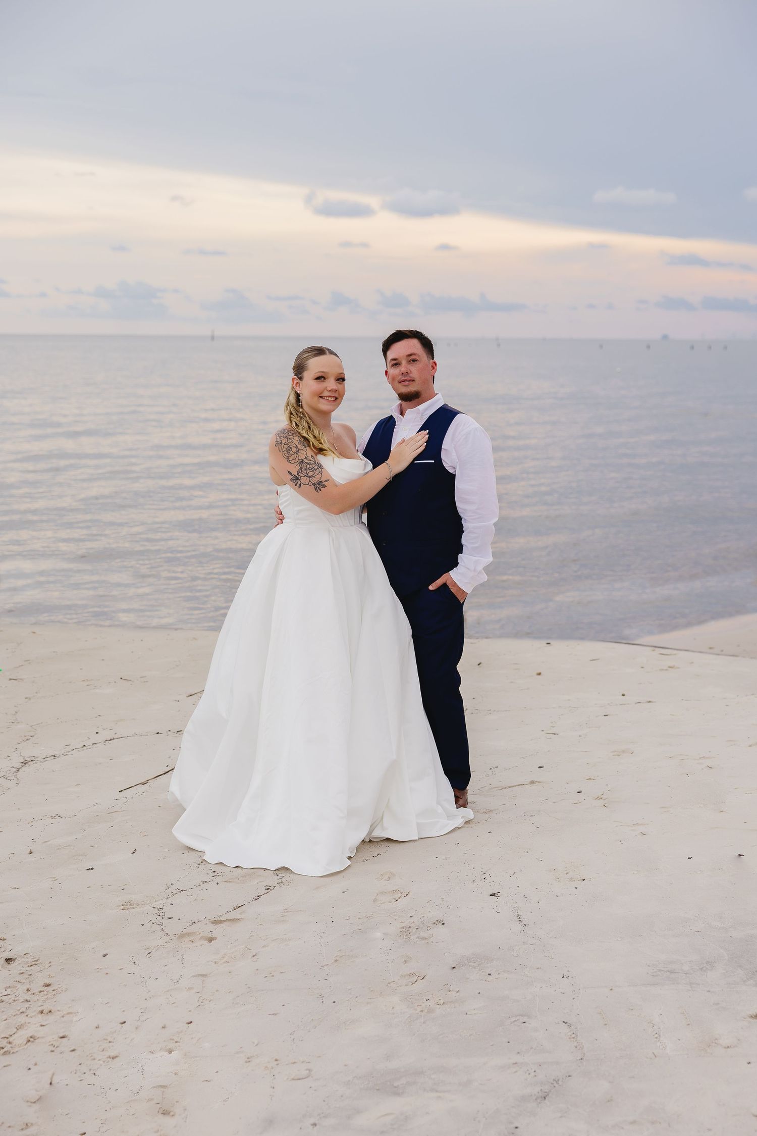 Newlywed couple embracing on white sandy beach at sunset with calm ocean waters in the background.