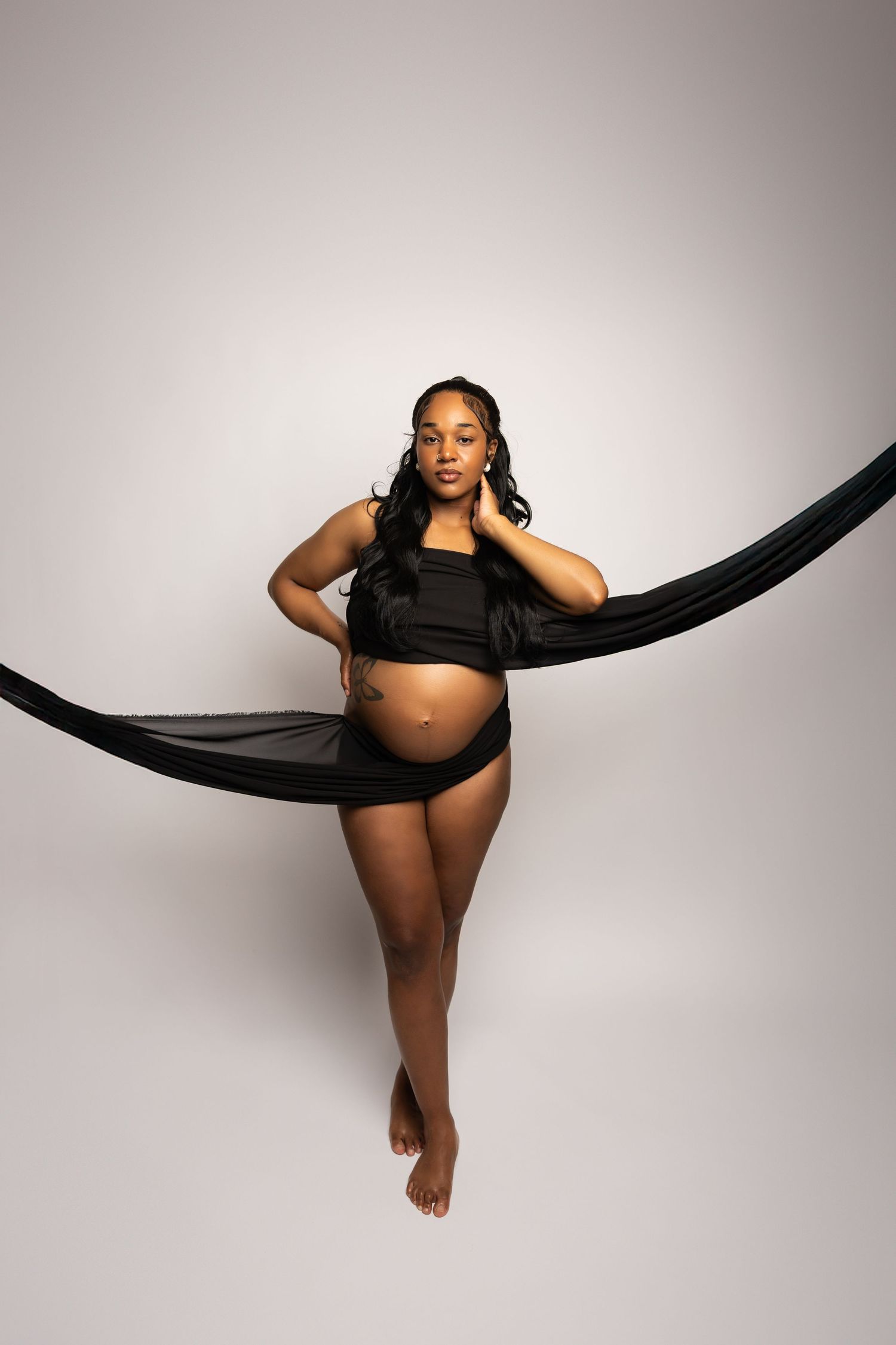 Pregnant woman poses confidently with black fabric draped around her in studio setting.