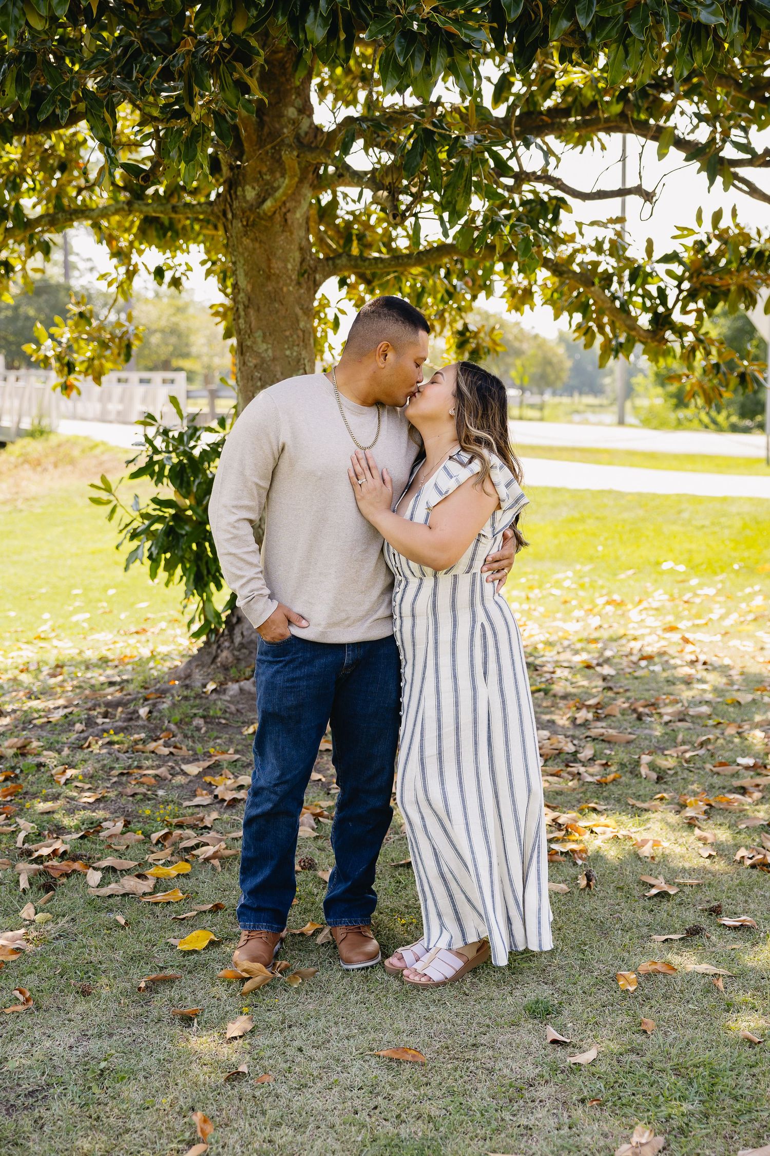 Couple sharing a romantic kiss under a large tree in a sunlit park.