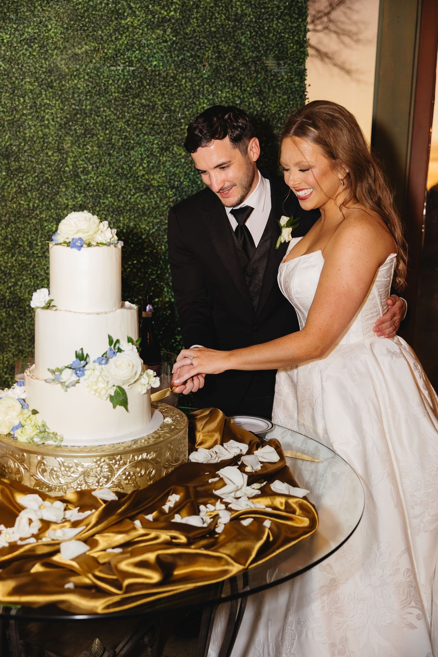 Smiling bride and groom cut a three-tiered white wedding cake decorated with blue and white flowers.