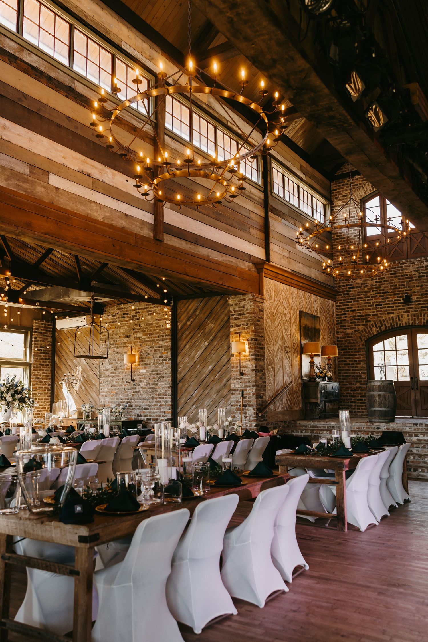Rustic wedding reception venue with exposed brick, wood beams, chandeliers, and elegantly set long tables.