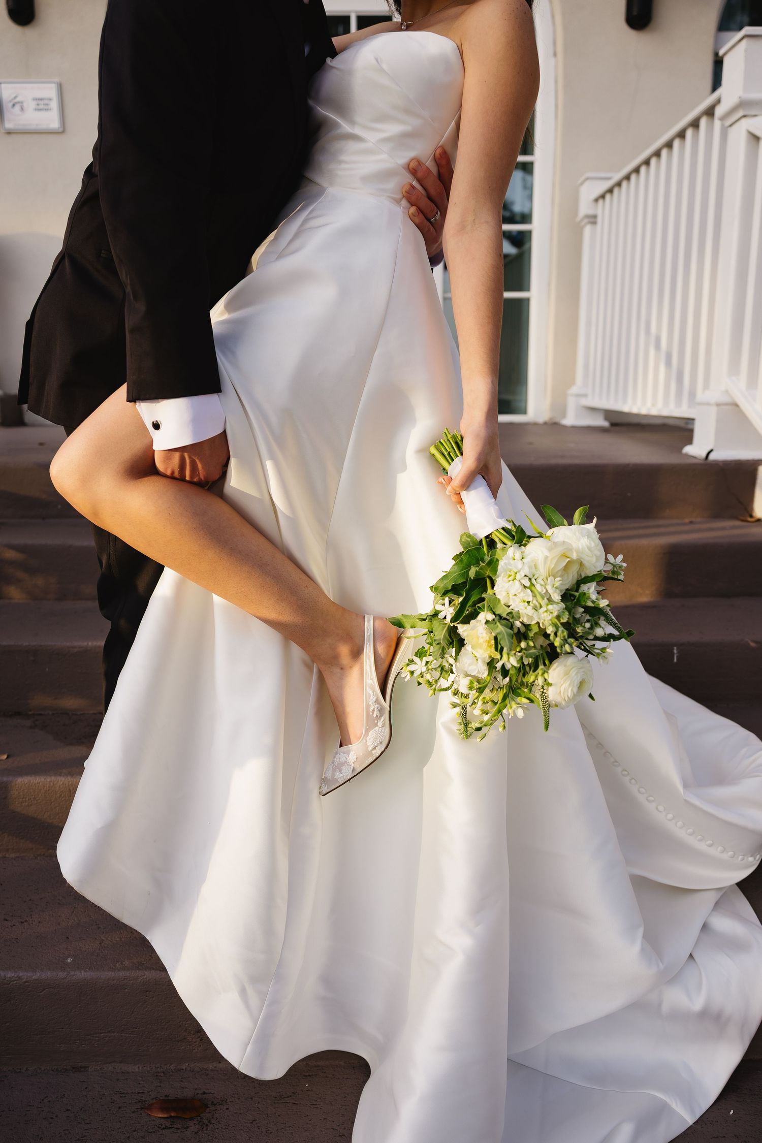 Close-up of a bride in a white strapless gown holding a white bouquet while being lifted by groom.