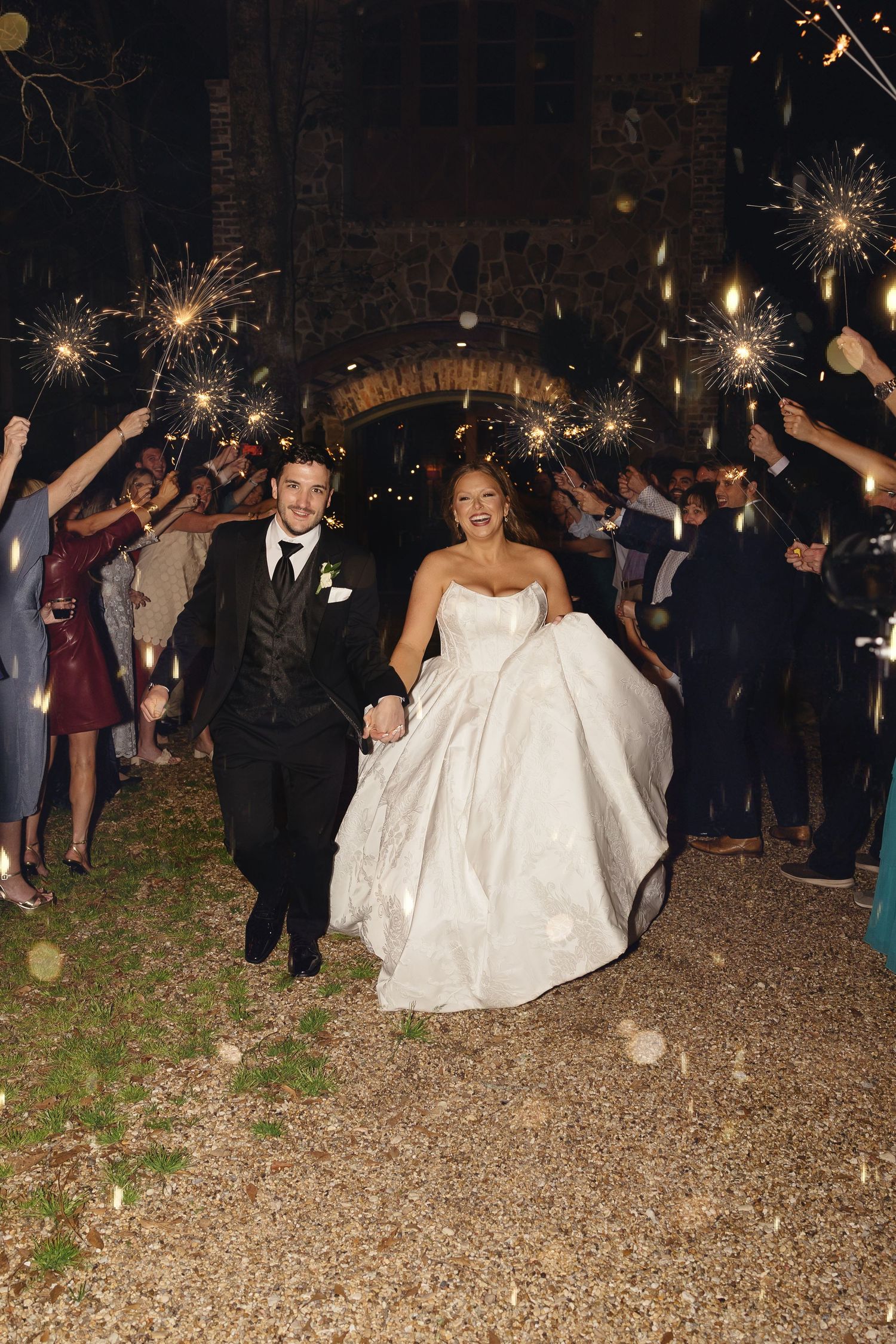 Joyful newlyweds exit through sparkler tunnel at night wedding celebration with cheering guests.