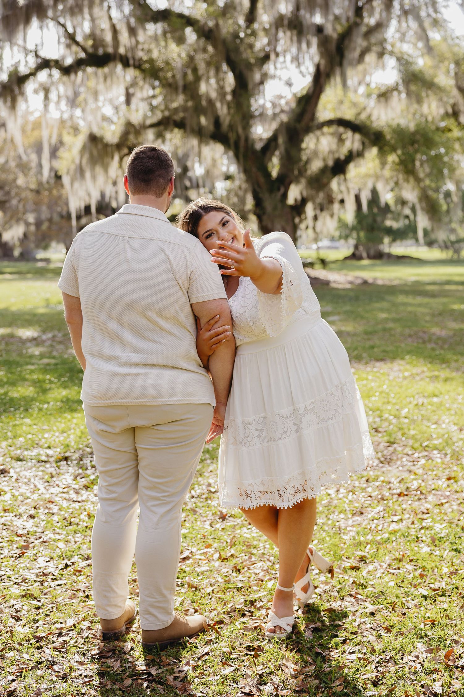 Woman in white dress hugs mans arm while showing engagement ring to camera outdoors.