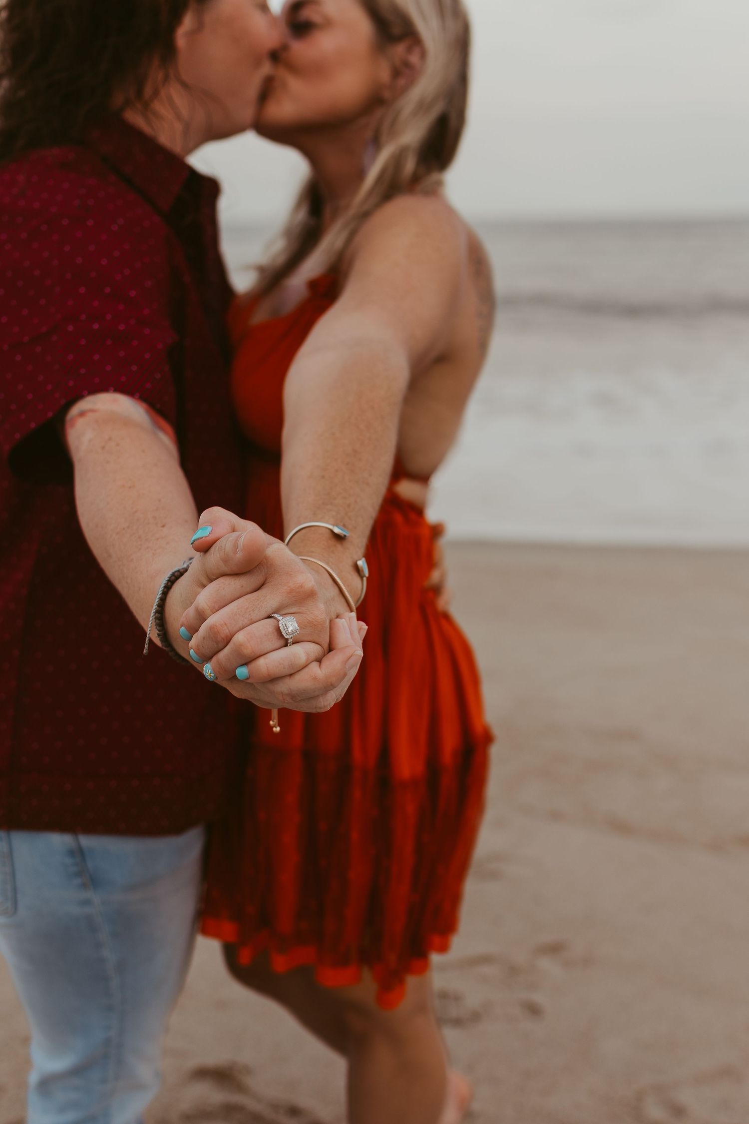 Newly engaged couple kisses on beach while showing off diamond engagement ring on woman's hand.