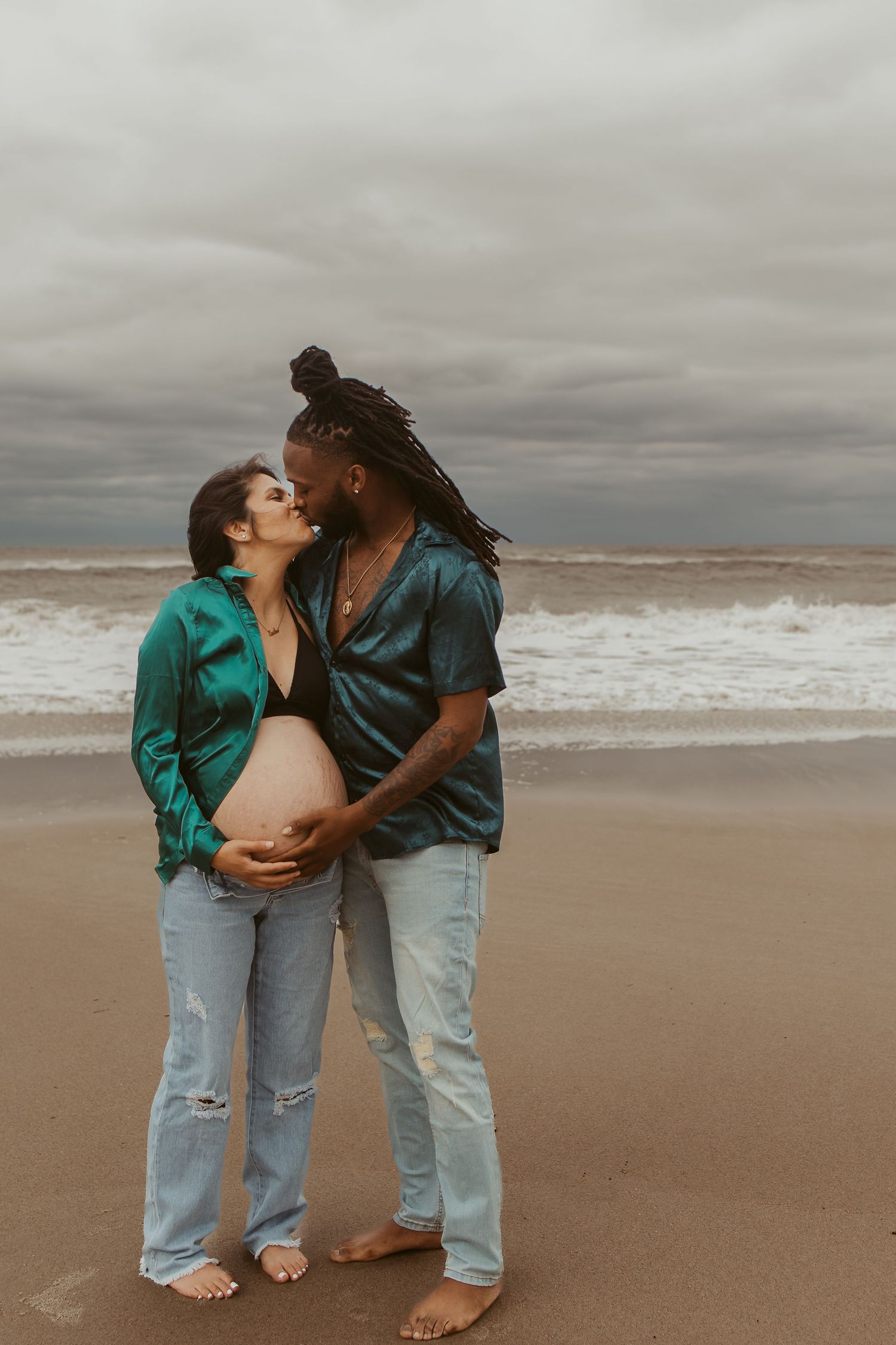 Interracial couple kissing on beach during maternity photoshoot, both wearing matching teal shirts.