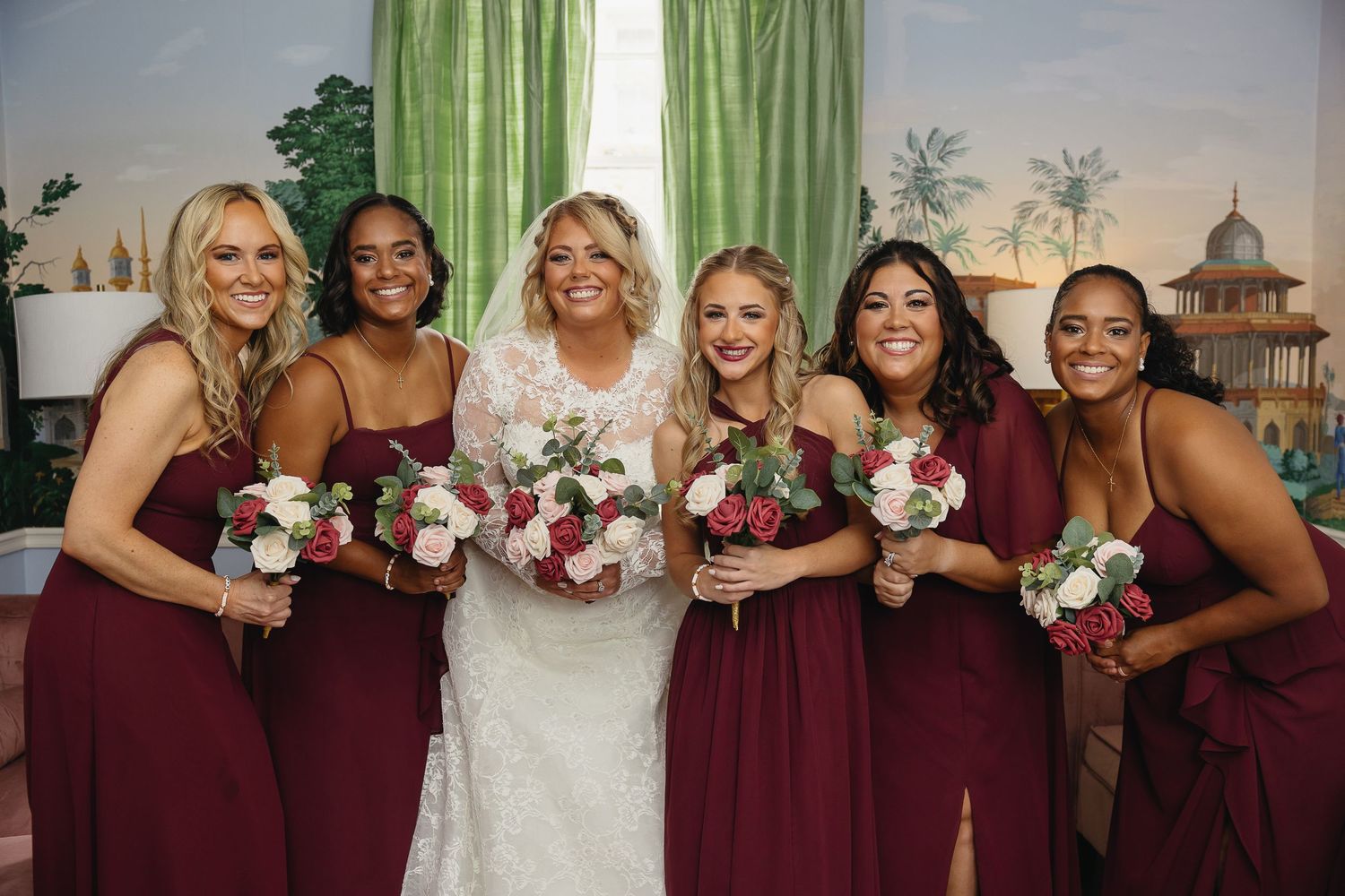 Bride in lace gown smiles with five bridesmaids in burgundy dresses holding floral bouquets.