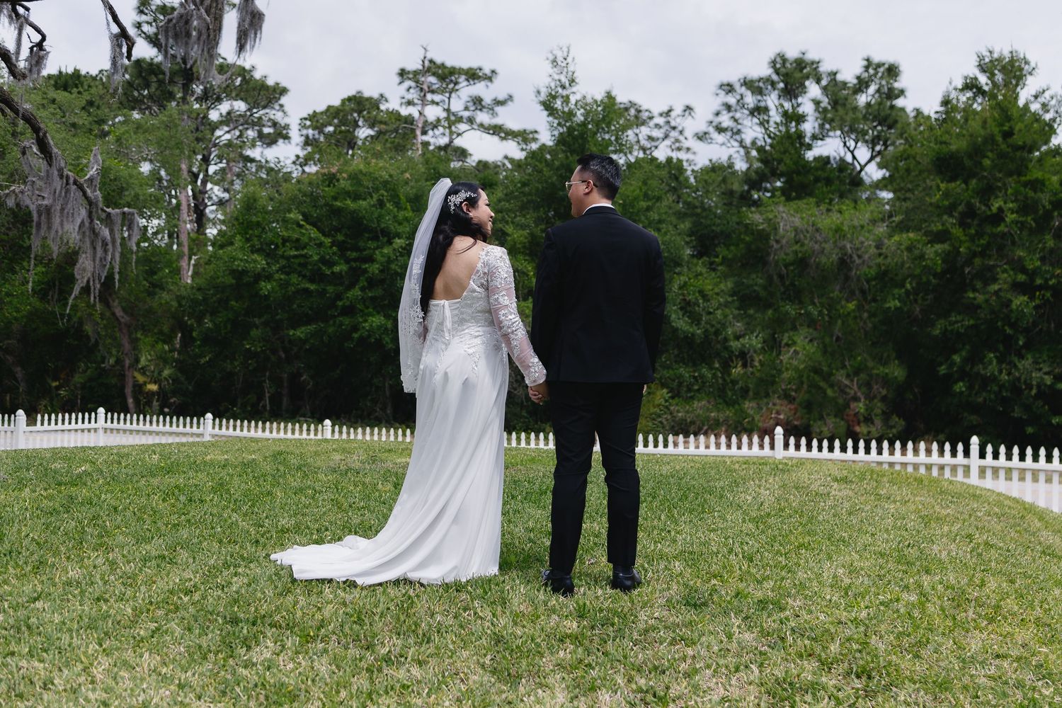 Bride and groom holding hands facing lush green trees in white fenced garden.