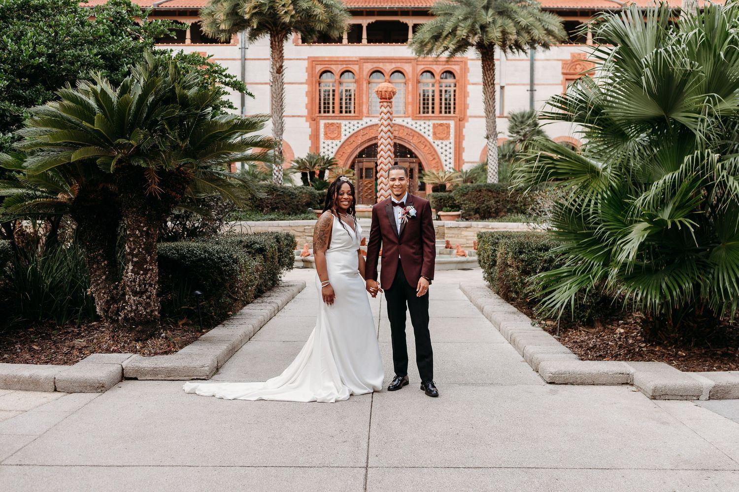 Interracial wedding couple holding hands in front of historic building surrounded by palm trees.