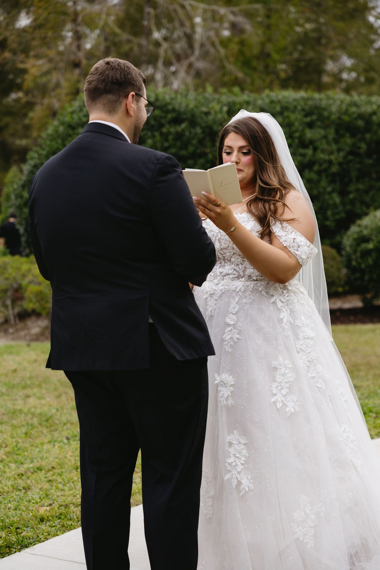 Bride in lace off-shoulder gown reads wedding vows from a card to groom in black suit outdoors.