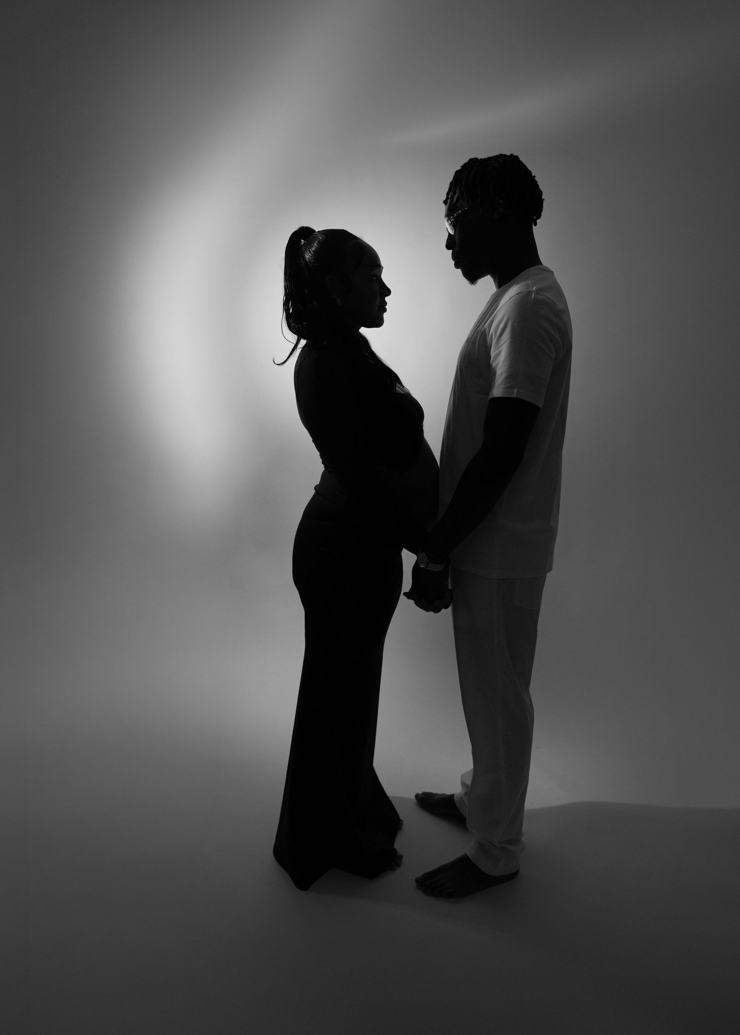 Black and white silhouette of pregnant couple holding hands facing each other in soft studio light.