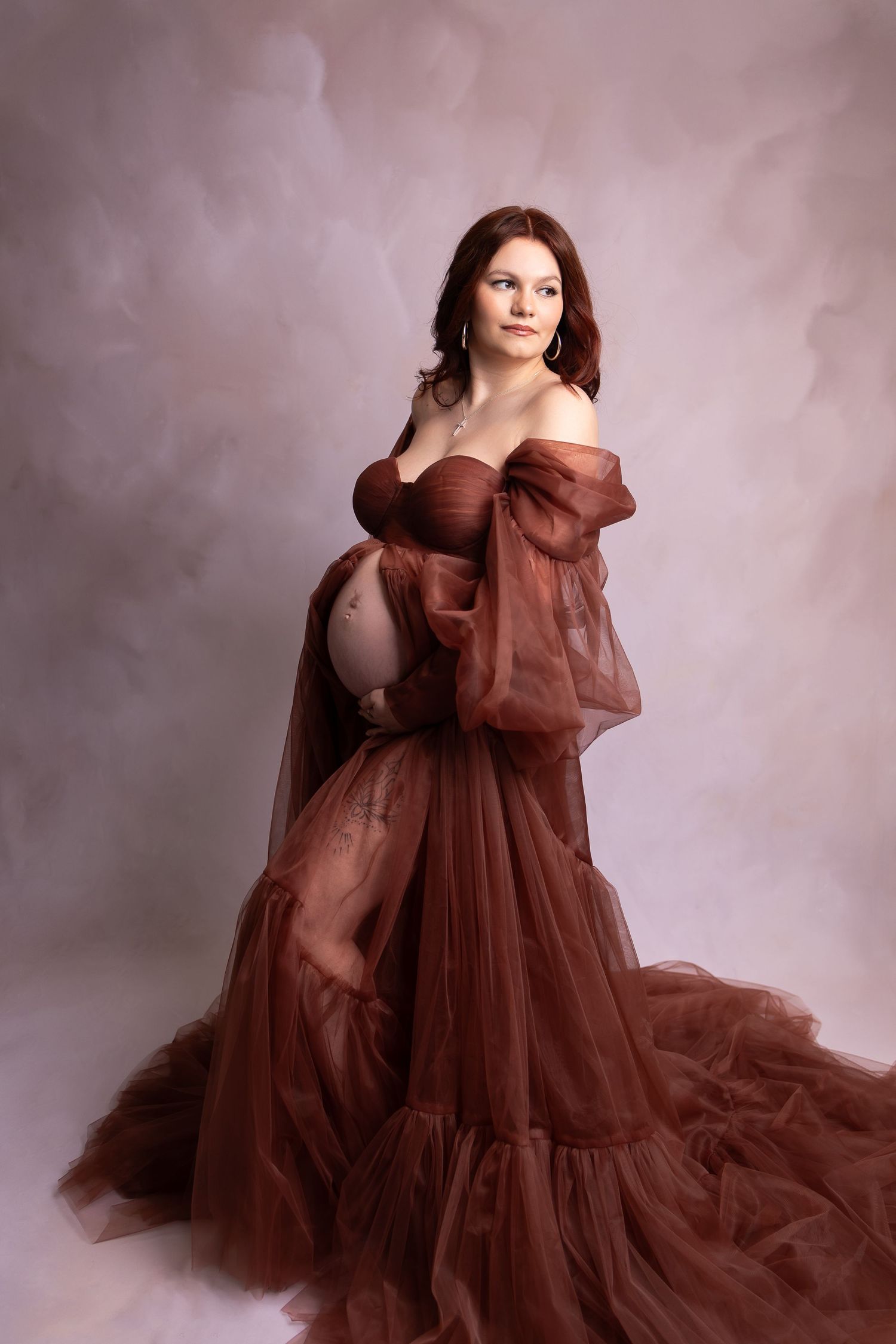 Elegant pregnant woman in an off-shoulder rust tulle maternity gown posing against a pink studio backdrop.