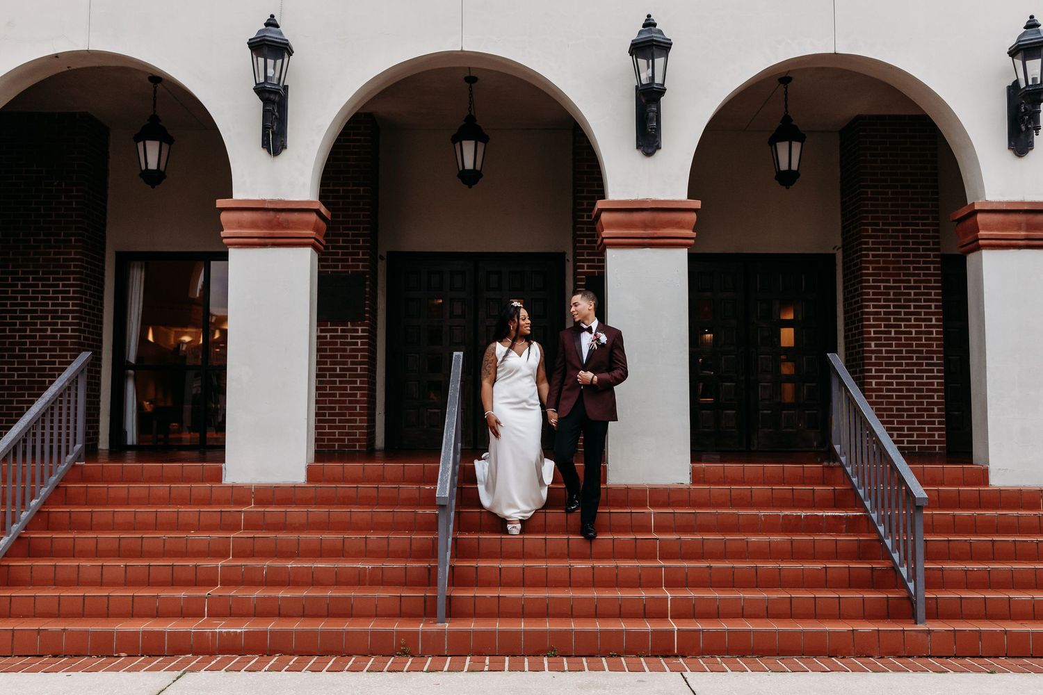 Wedding couple walking down brick steps of elegant building with arched columns and lanterns.