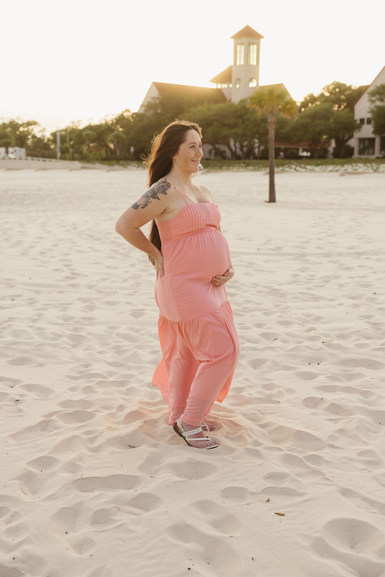 Glowing pregnant woman in pink dress smiling on sunny beach with resort buildings behind her.
