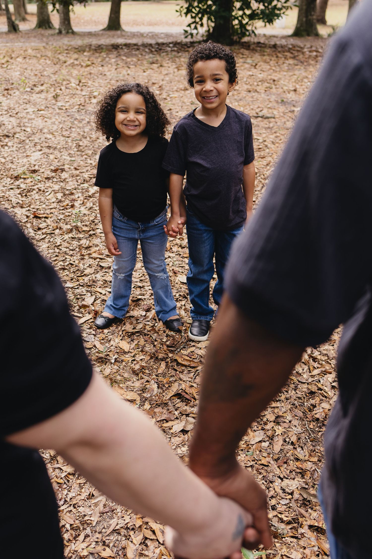 Two young children smiling and holding hands with parents in a leaf-covered park setting.