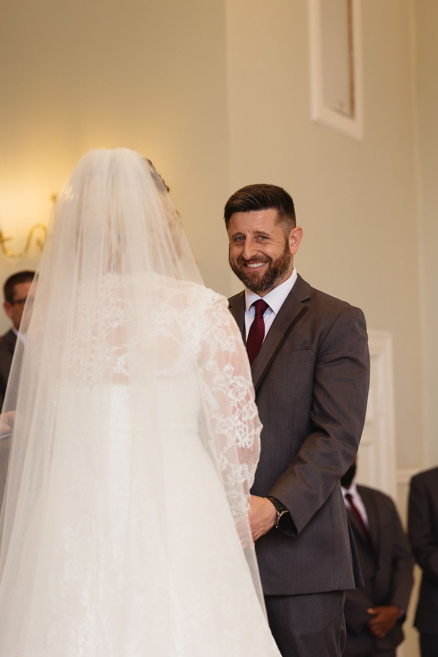 Groom in gray suit smiling lovingly at bride in lace wedding dress during ceremony.