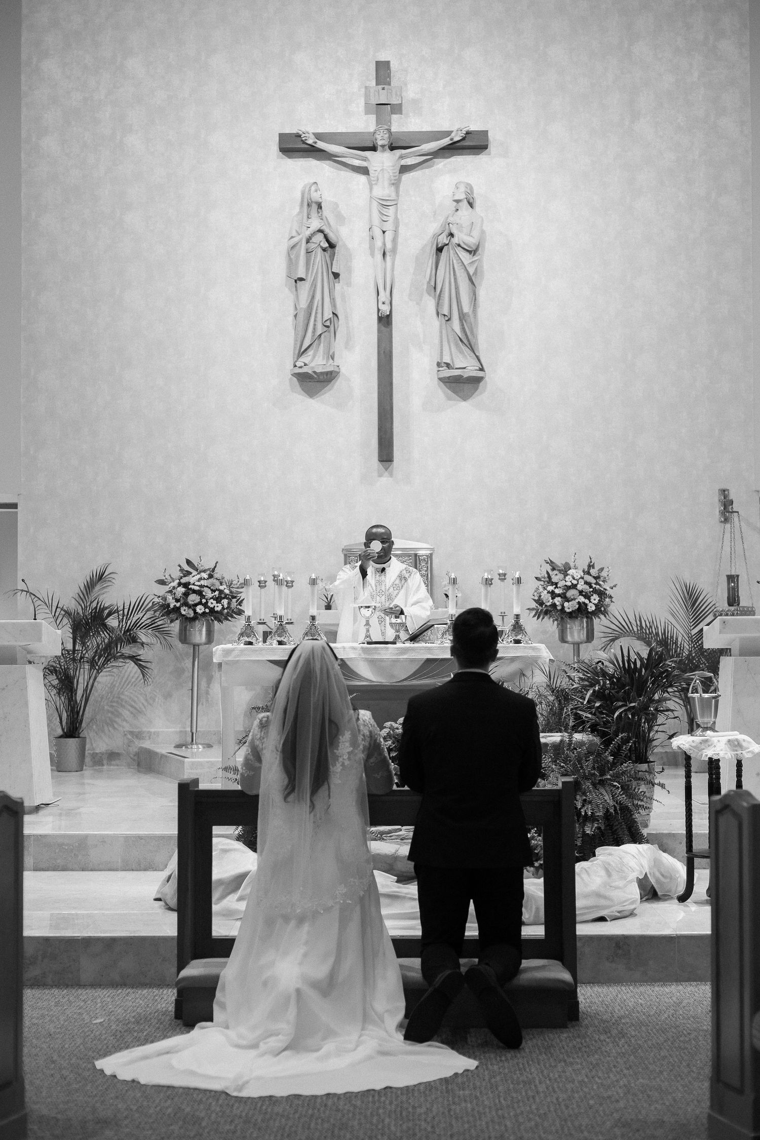 Black and white photo of bride and groom kneeling before priest at church altar.