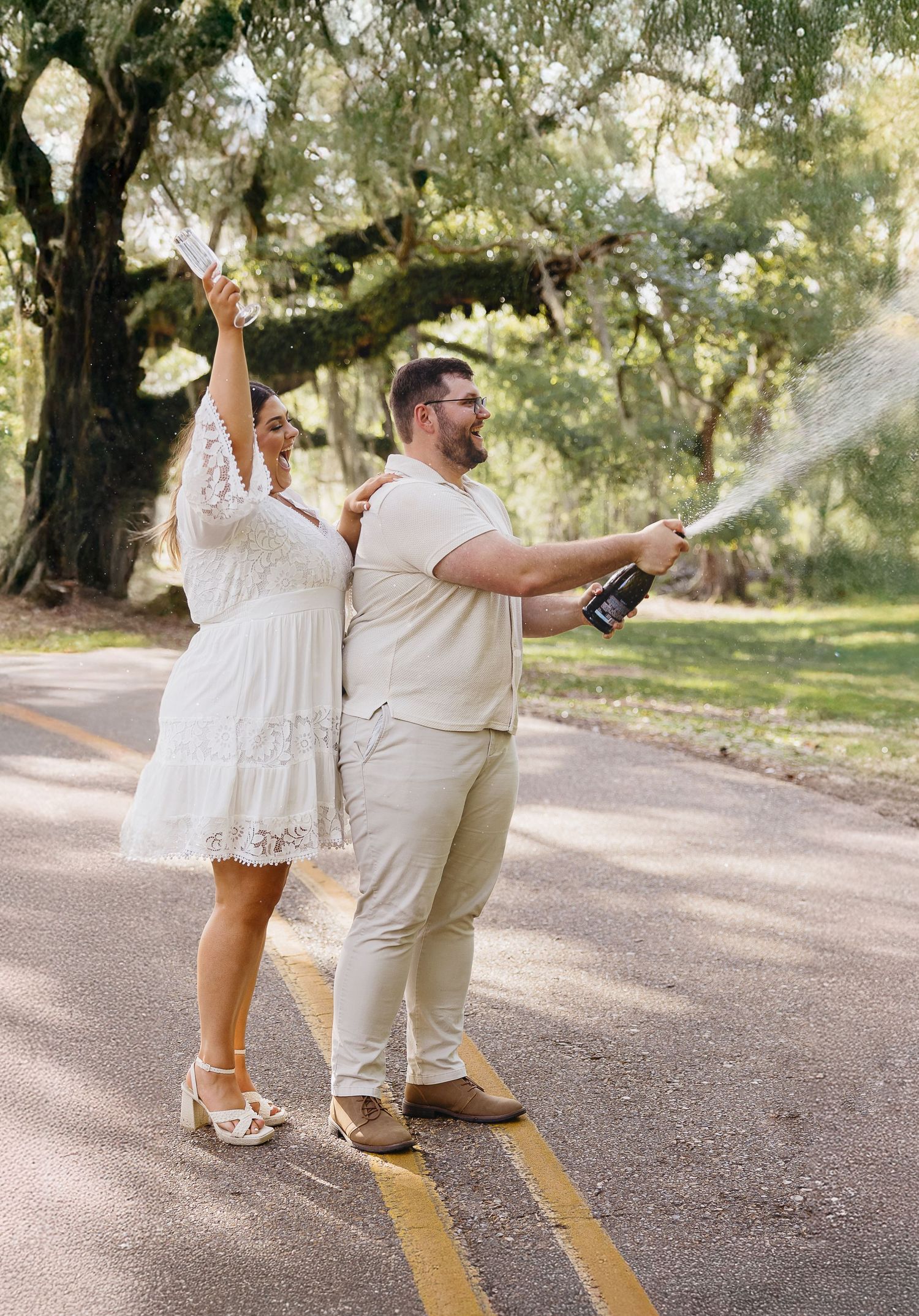 Joyful couple popping champagne on a road, woman in white lace dress raising paper while man sprays bottle.