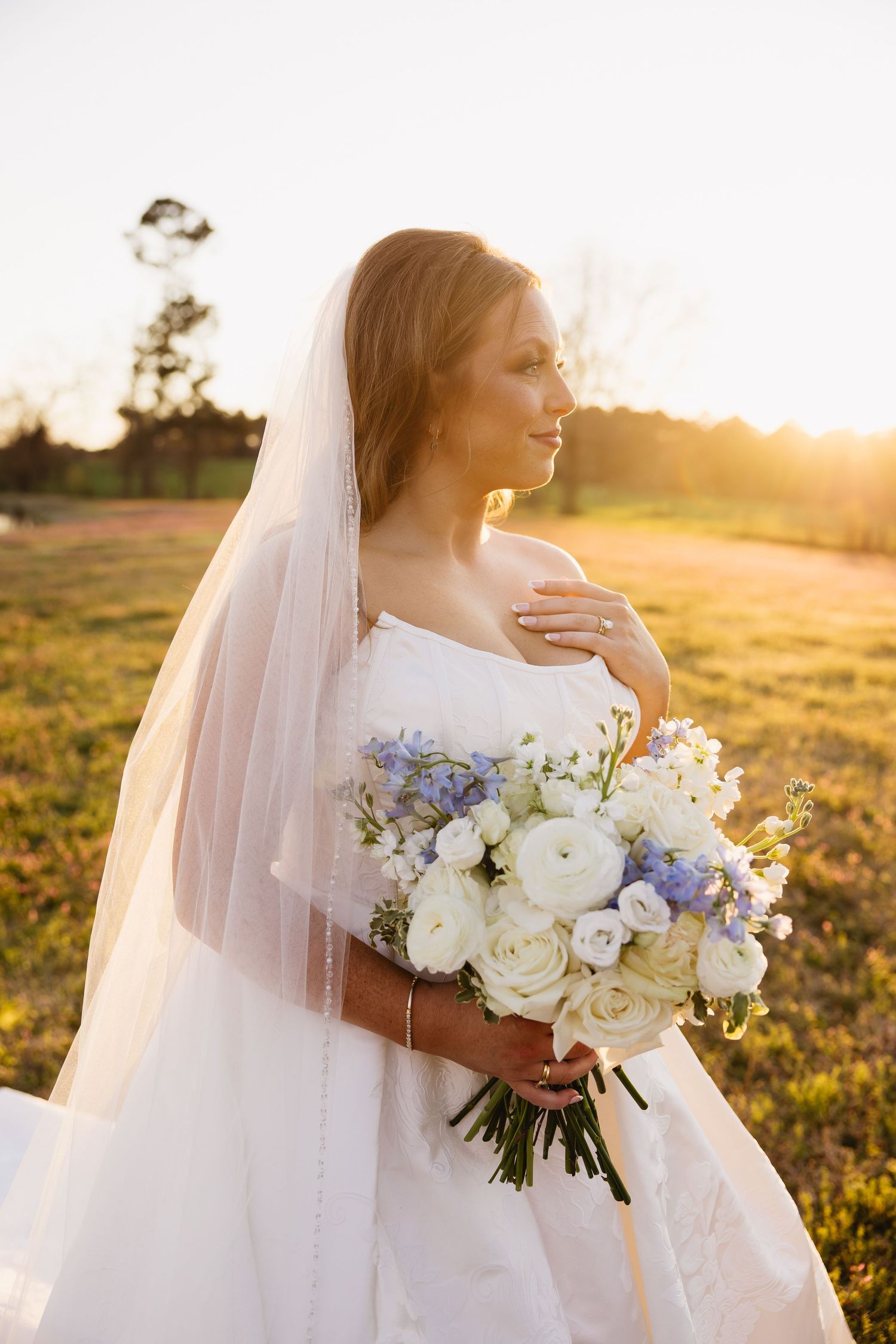 Bride in white off-shoulder gown holding a bouquet of white and blue flowers at golden hour outdoors.