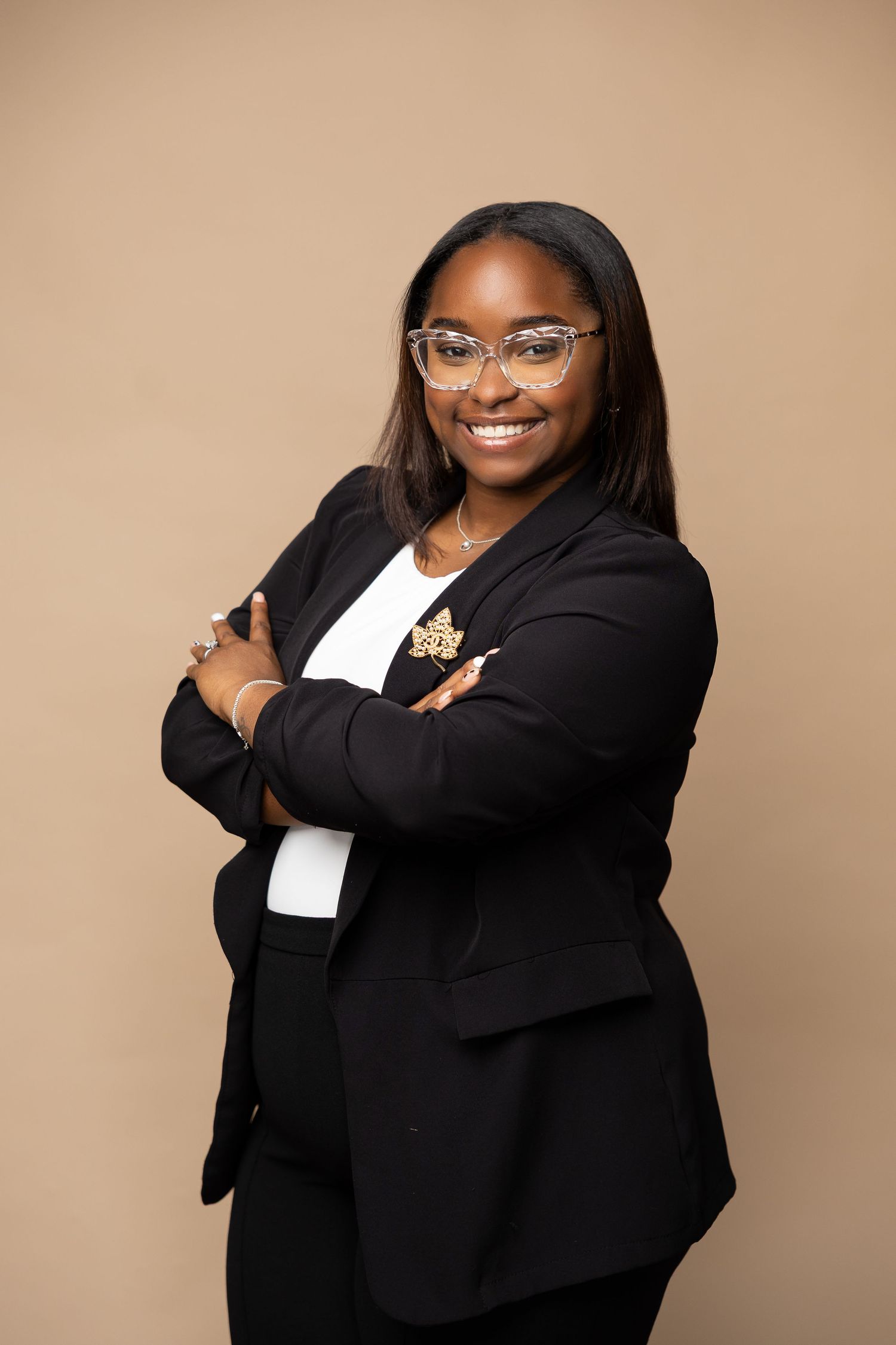 Professional Black woman in black blazer smiles with arms crossed wearing glasses on beige background.