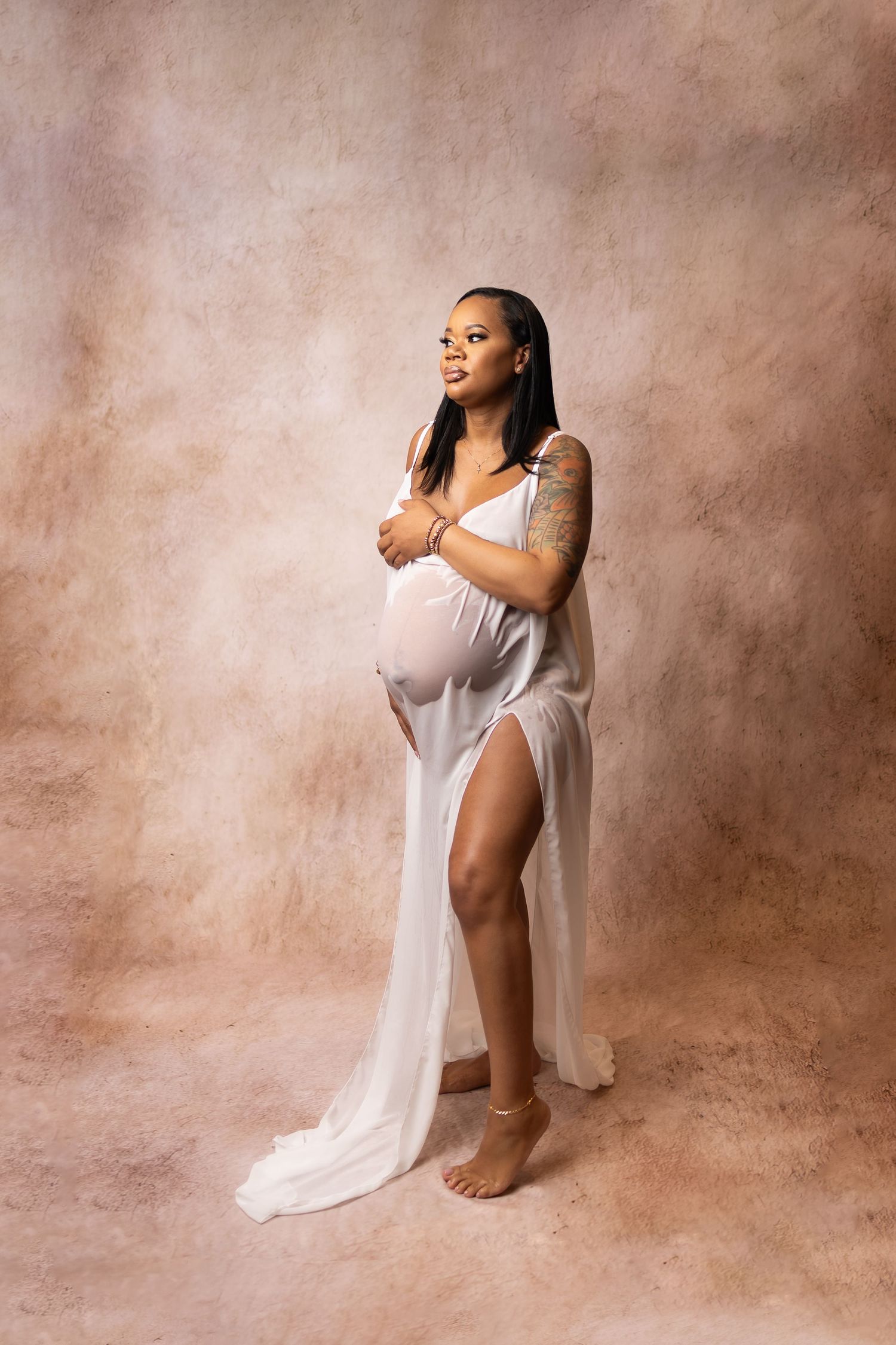 Smiling pregnant woman in sheer white draped gown with tattoos poses in studio maternity shoot.