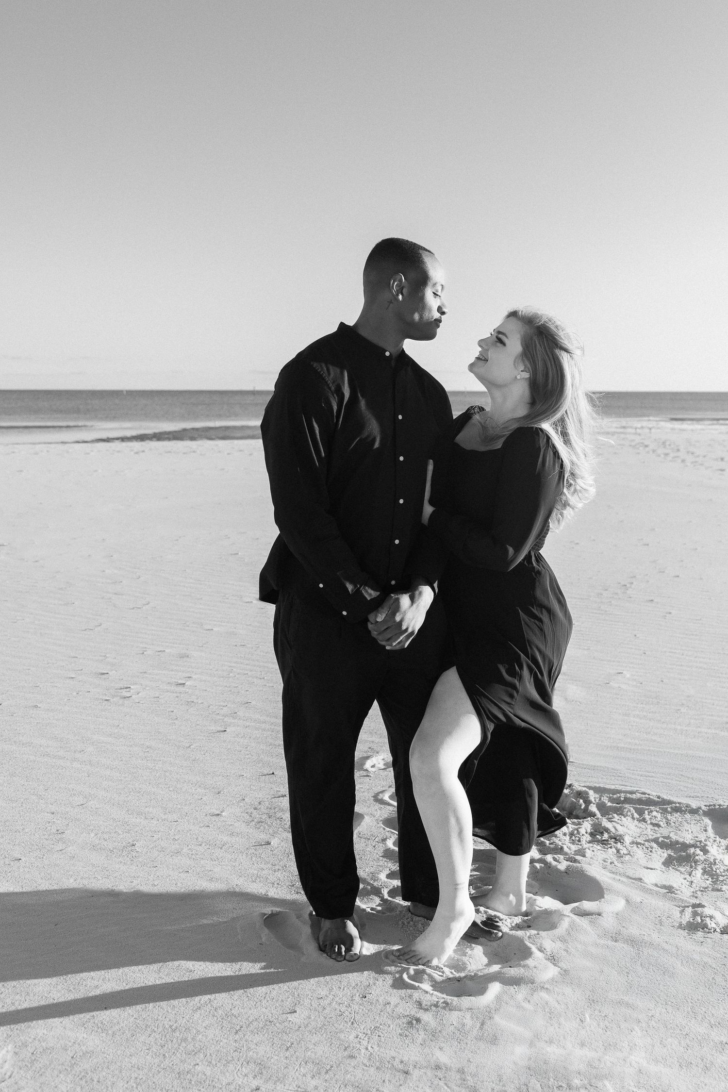 Black and white portrait of an interracial couple embracing romantically on a beach.